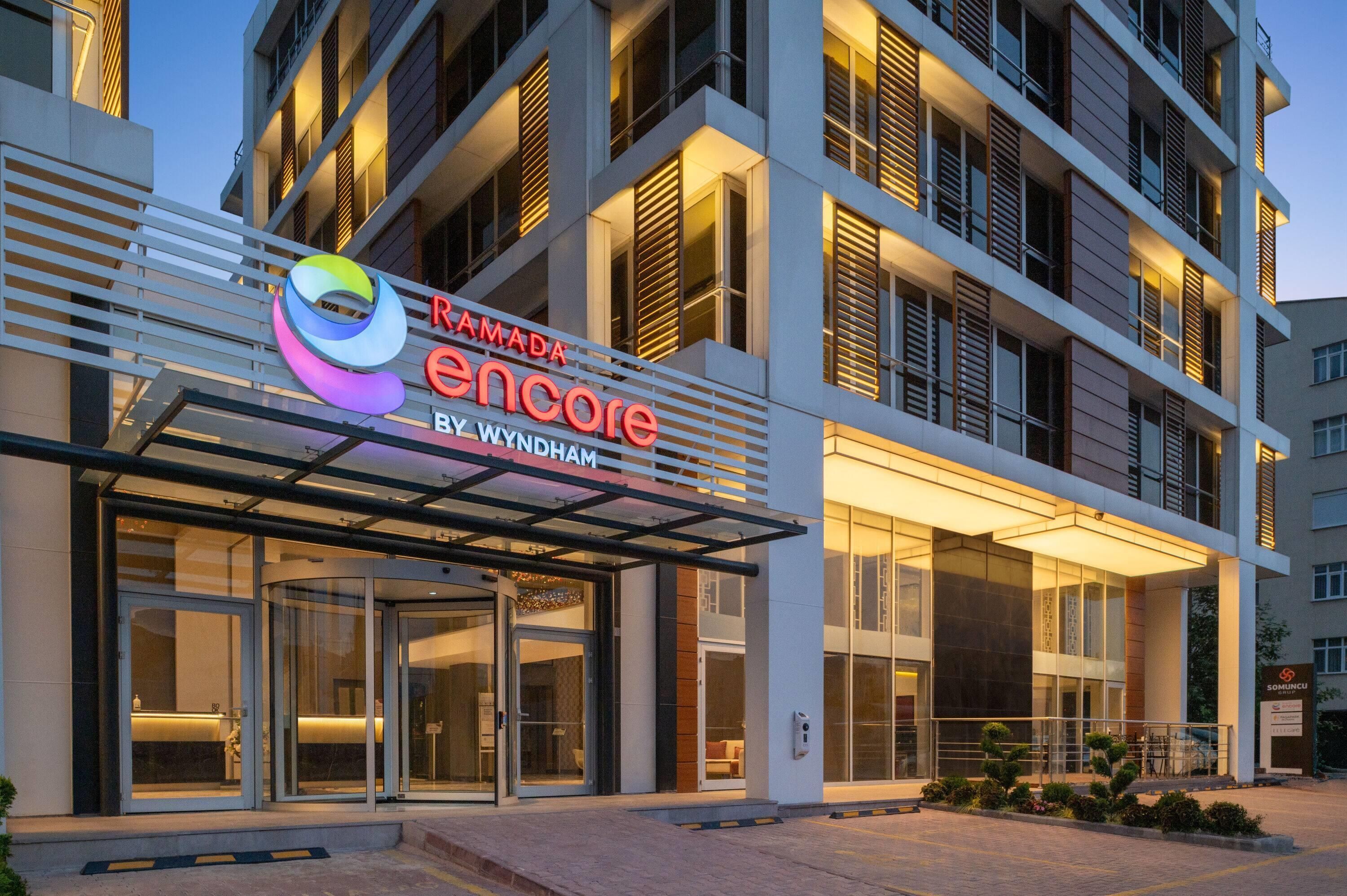 A modern hotel entrance with a glass facade, a prominent sign for Ramada Encore, and a well-lit exterior.