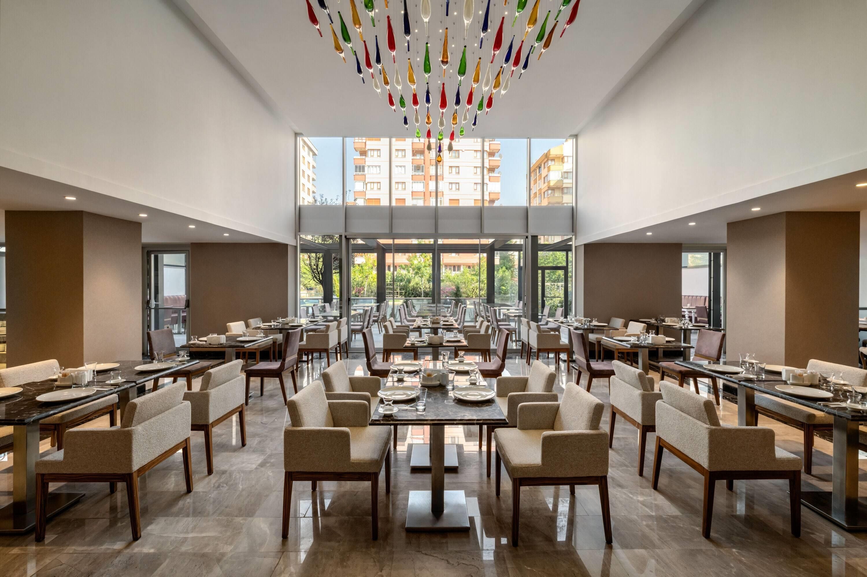 A spacious dining area with multiple tables and chairs, large windows offering a view of the cityscape, and a modern chandelier.