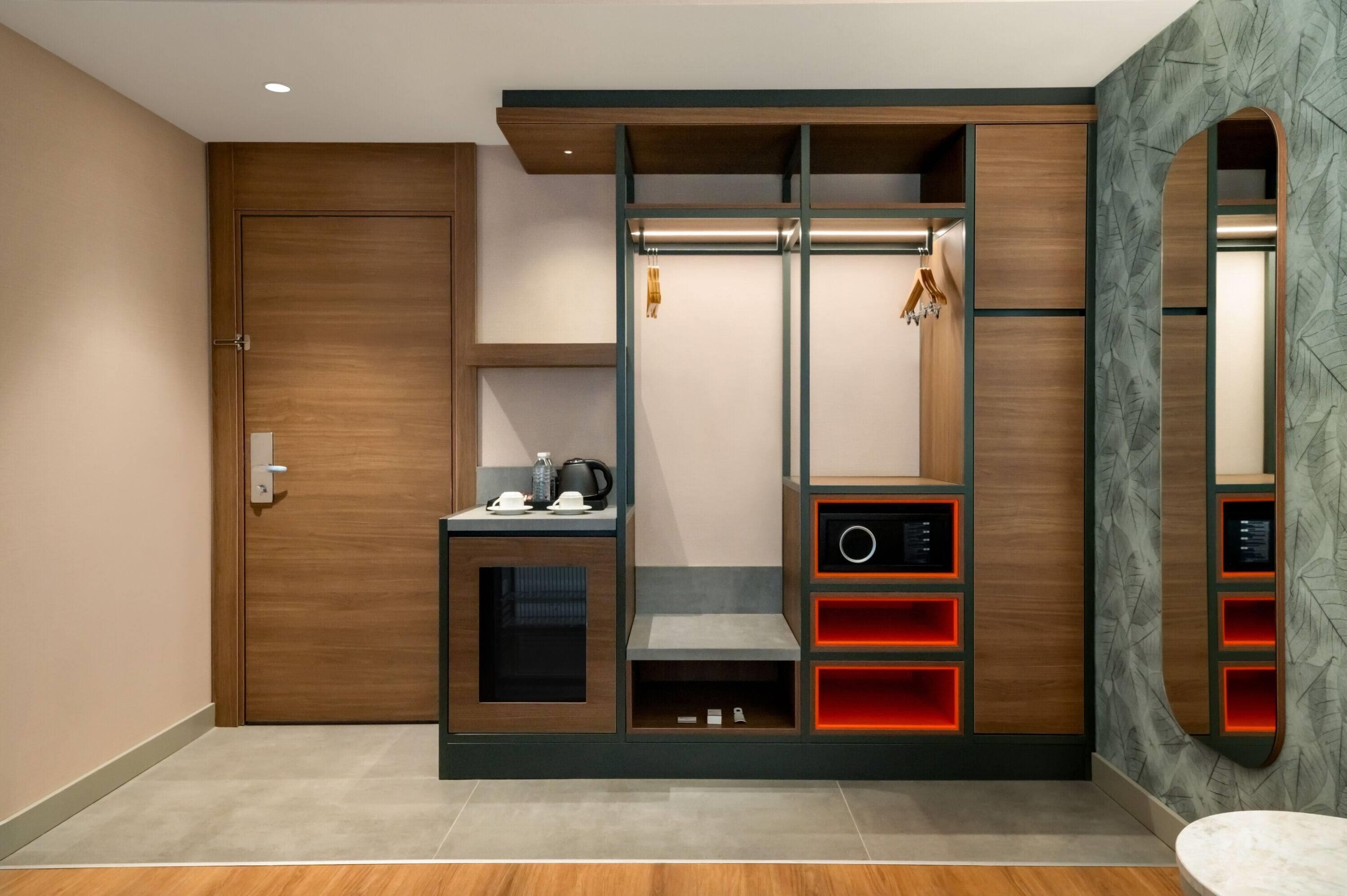 Una habitación moderna con un armario de madera, una pequeña zona de estar con una mesa, y un espejo con un papel tapiz estampado con hojas.