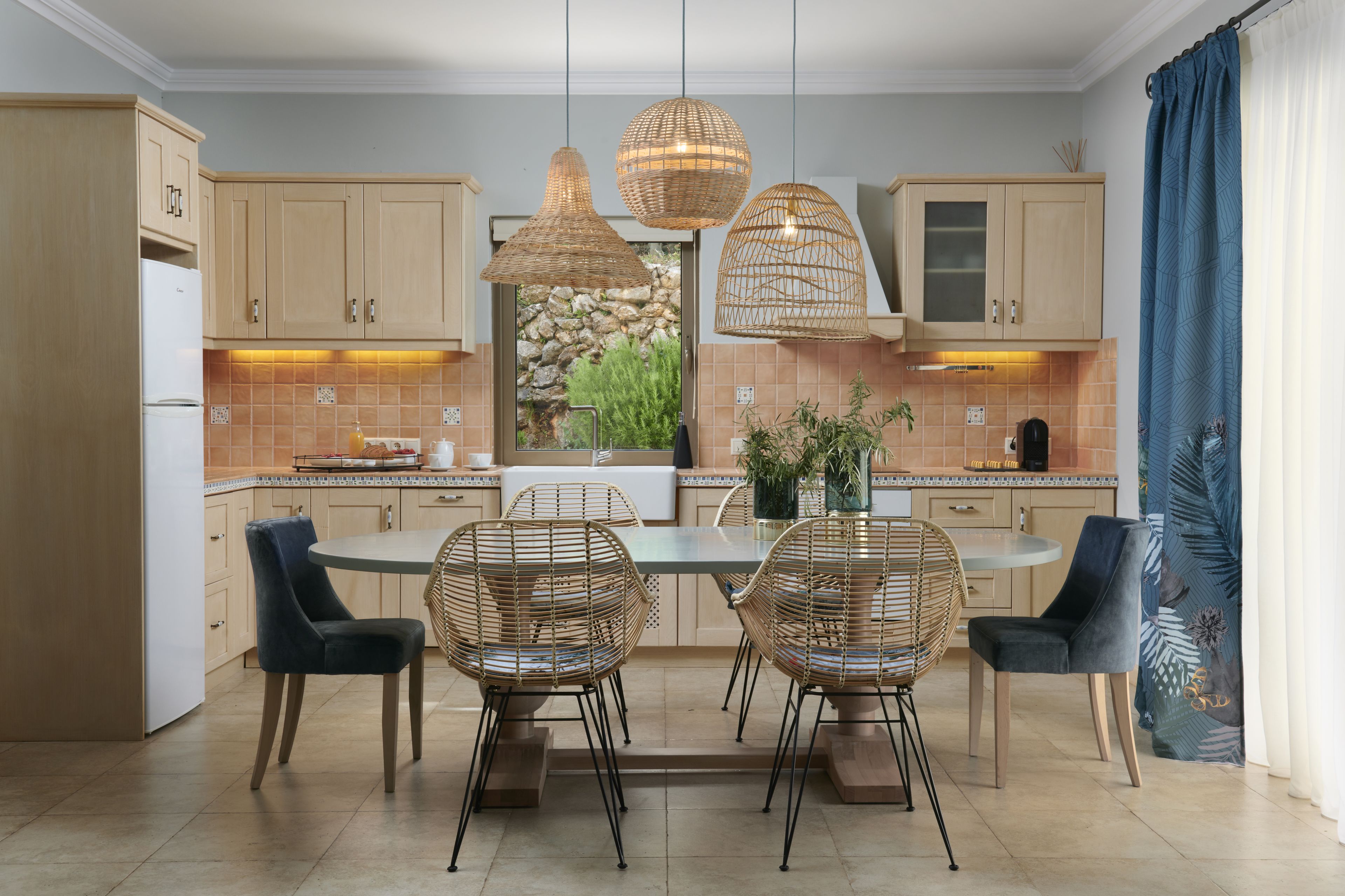 A modern kitchen with wooden cabinets, a central island with chairs, pendant lights, and a view of greenery outside.