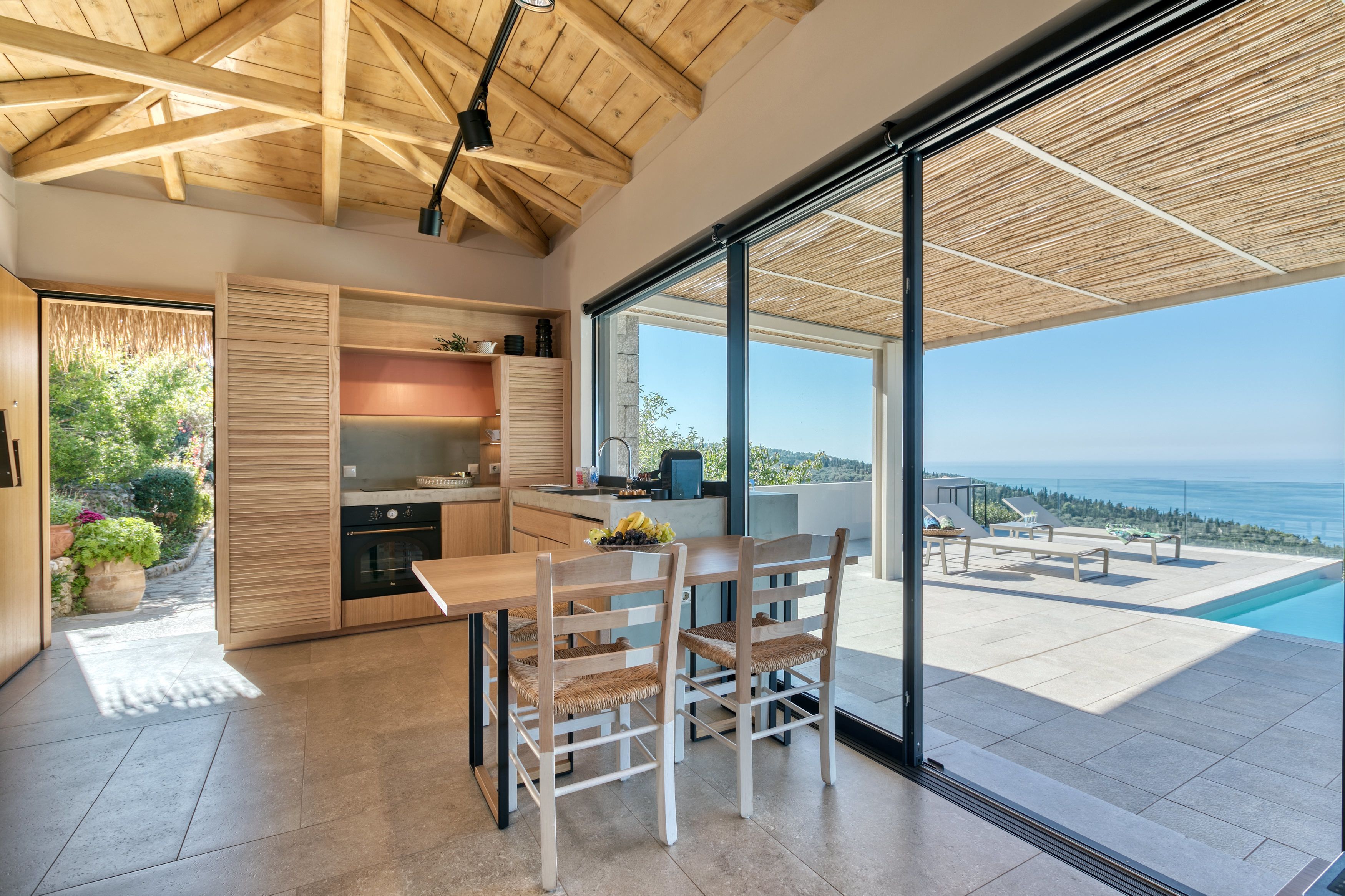 A modern kitchen with wooden beams, a dining area with a table and chairs, and a large sliding glass door leading to a balcony with a pool and ocean view.
