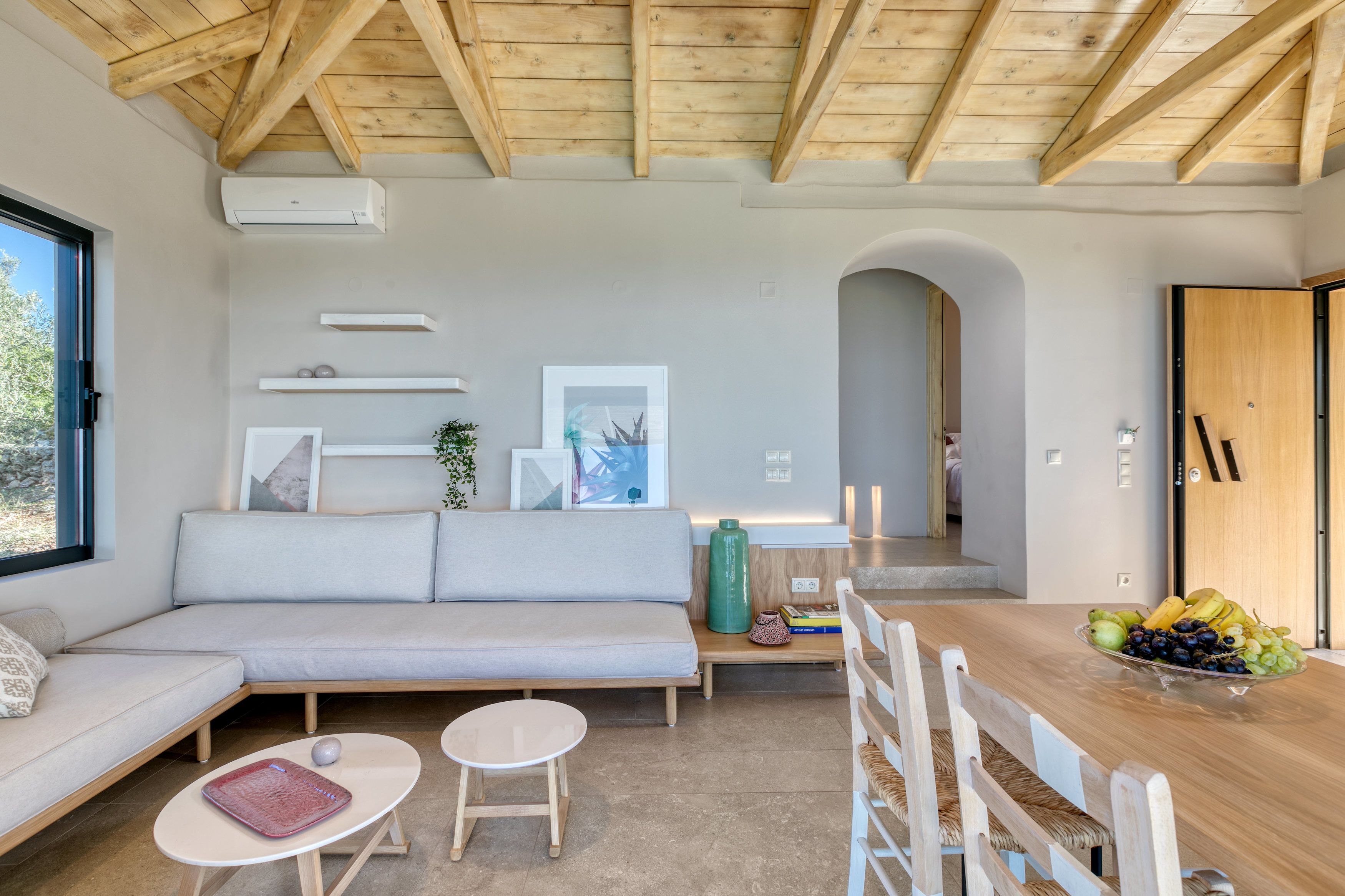 Un interior modern amb sostre de fusta, un sofà, una taula de dinar plena de fruita i una vista per la finestra.