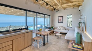 A modern kitchen with large windows, a dining area, and a living space with a view of the ocean. - Art Blue Villas (Lefkada)