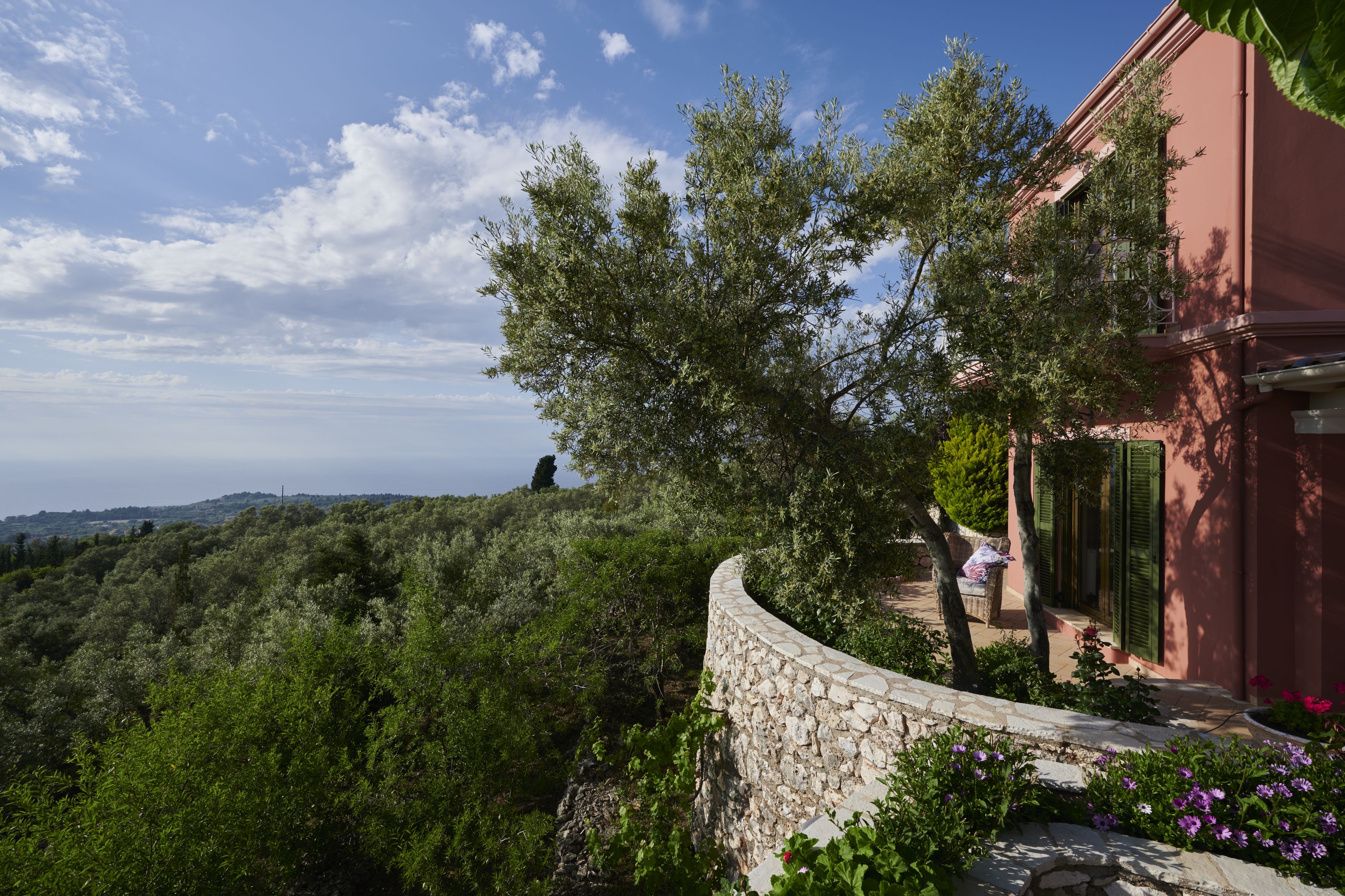 A stone wall with a curved top, a pink building with green shutters, and a view of a hilly landscape.