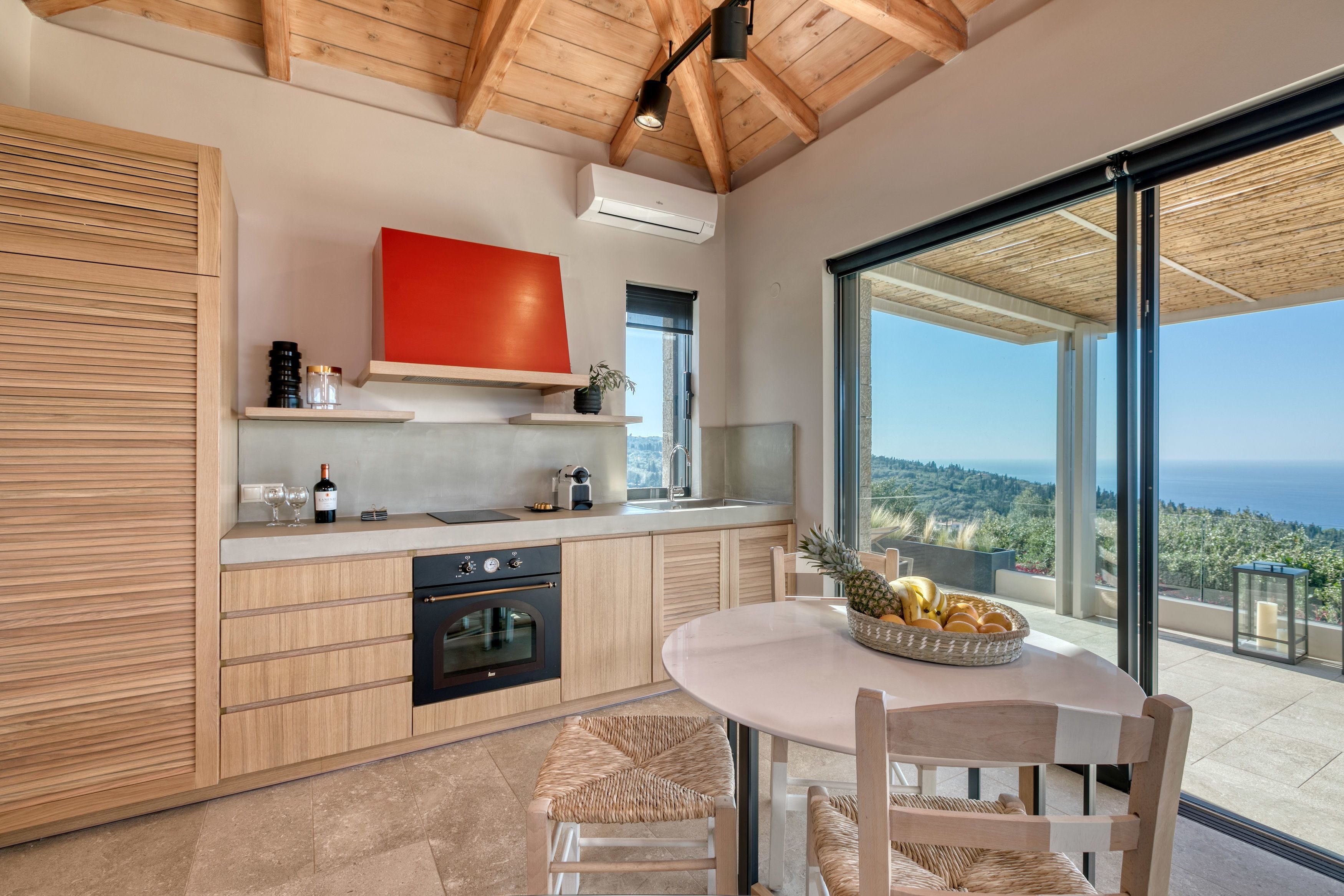 A modern kitchen with wooden cabinets, a red accent wall, and a dining area with a view of the sea.
