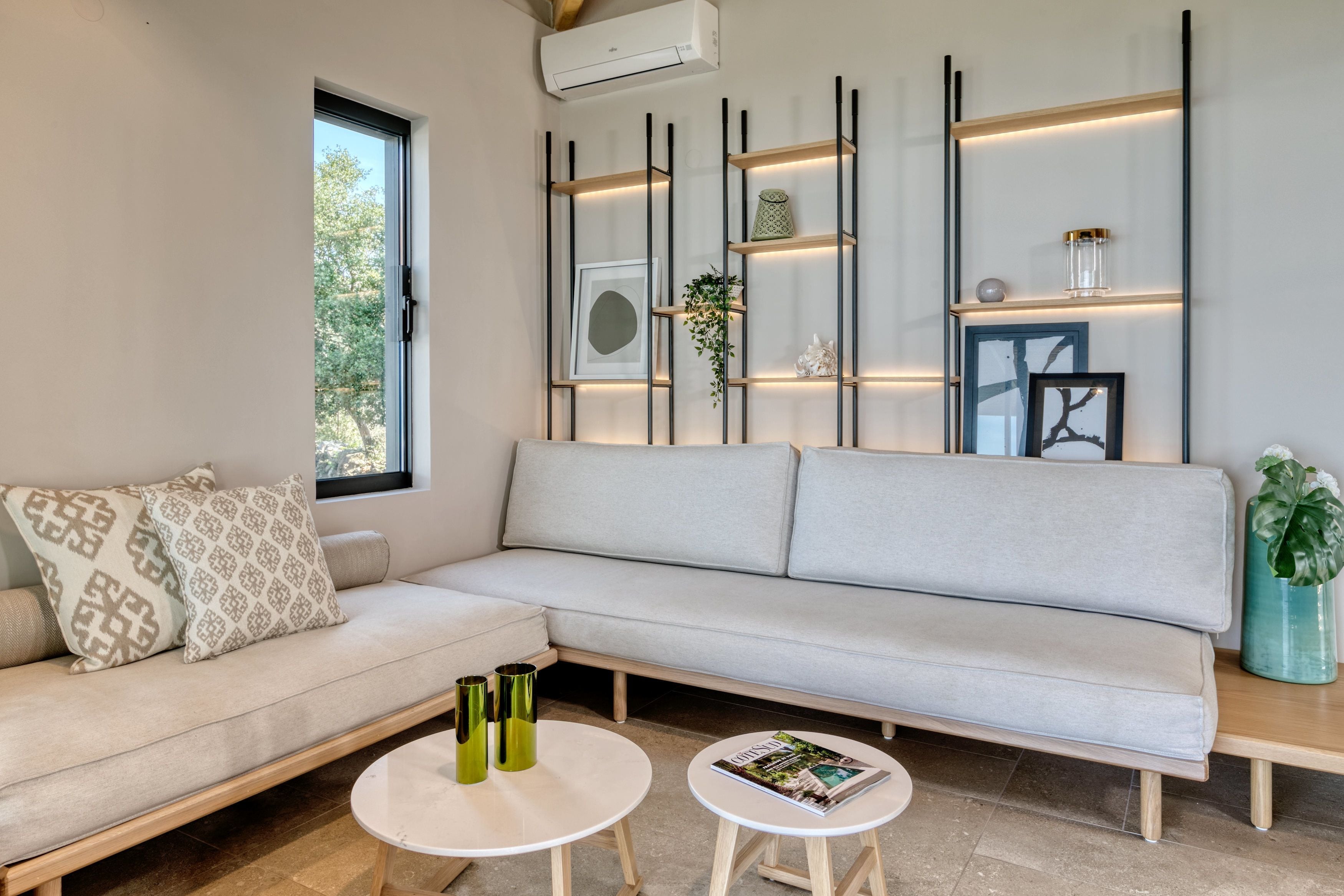 Una sala de estar moderna con un sofá de color claro, dos mesas redondas y un estante de pared con objetos decorativos.