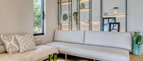 Una sala de estar moderna con un sofá de color claro, dos mesas redondas y un estante de pared con objetos decorativos.