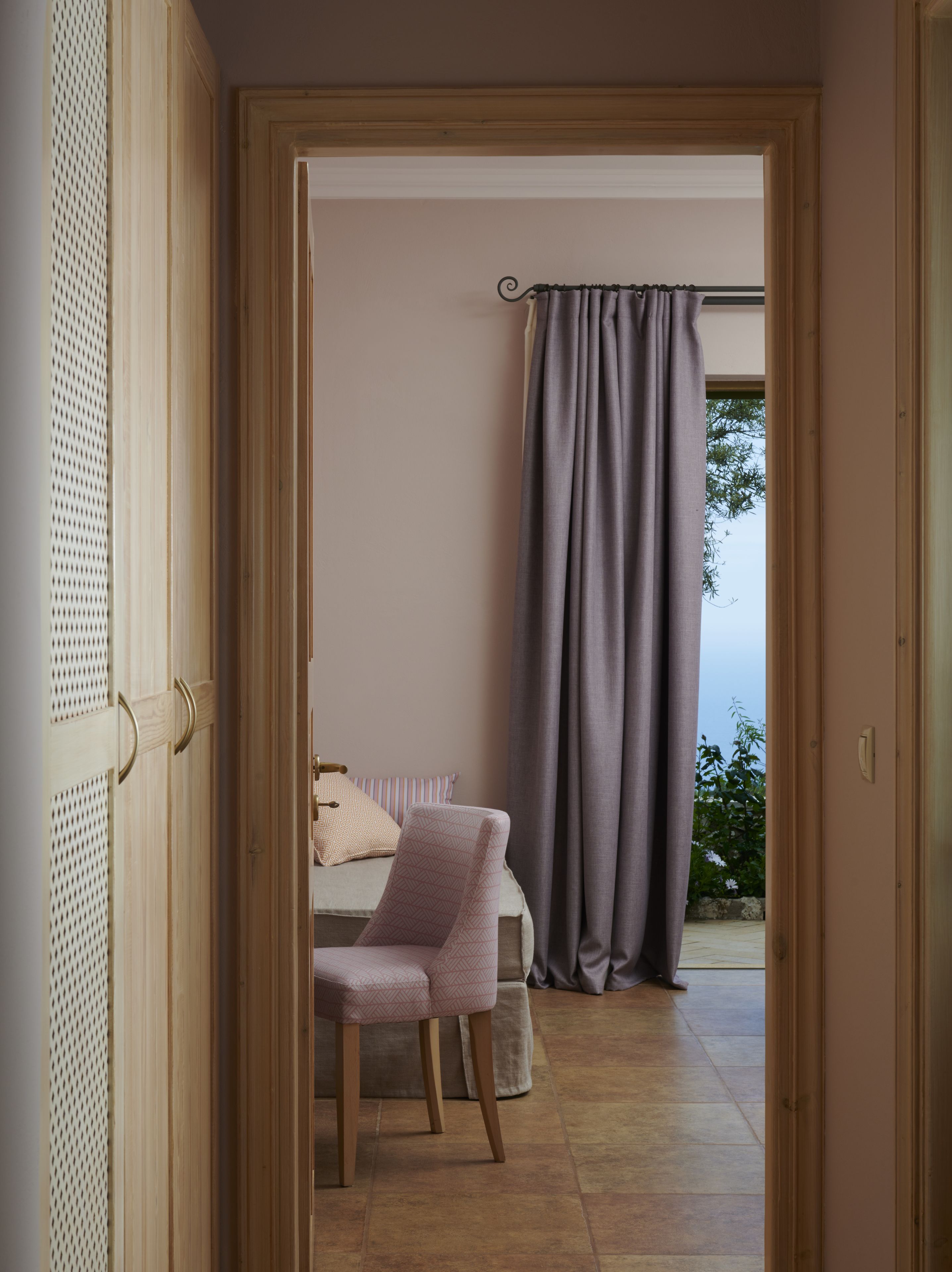 A room with a bed, a chair, and a view of the outdoors through a window with curtains.