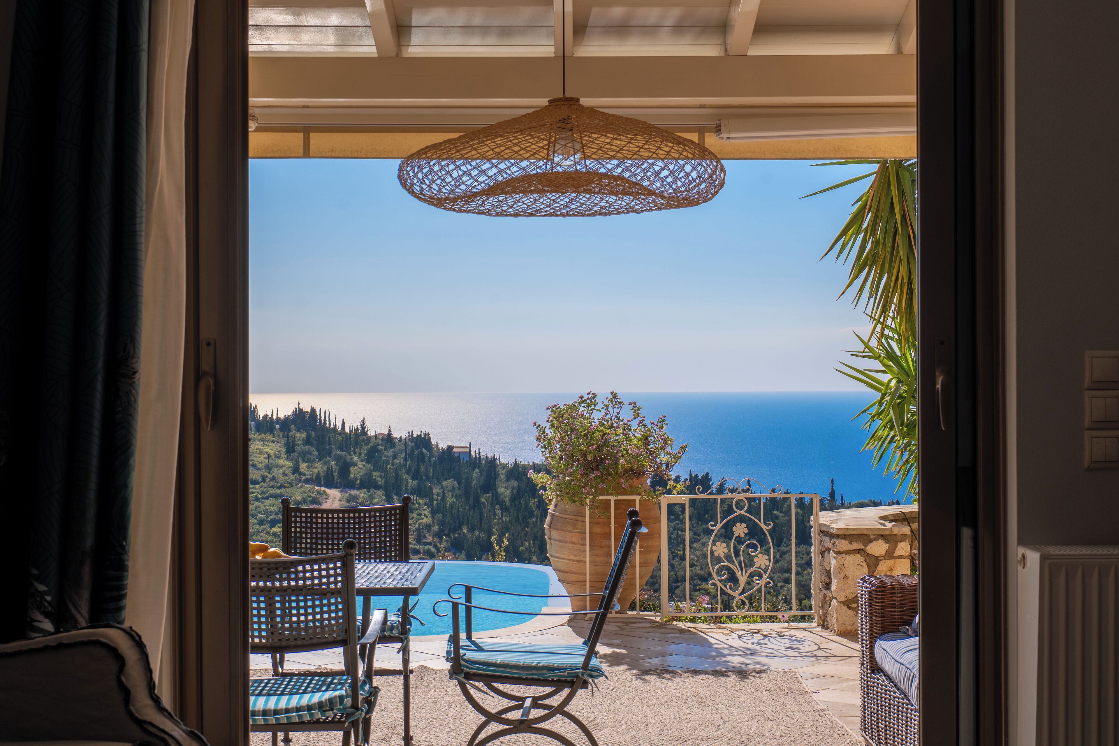 An open patio with a view of the sea, a wicker chair, a table, and a hanging light fixture.