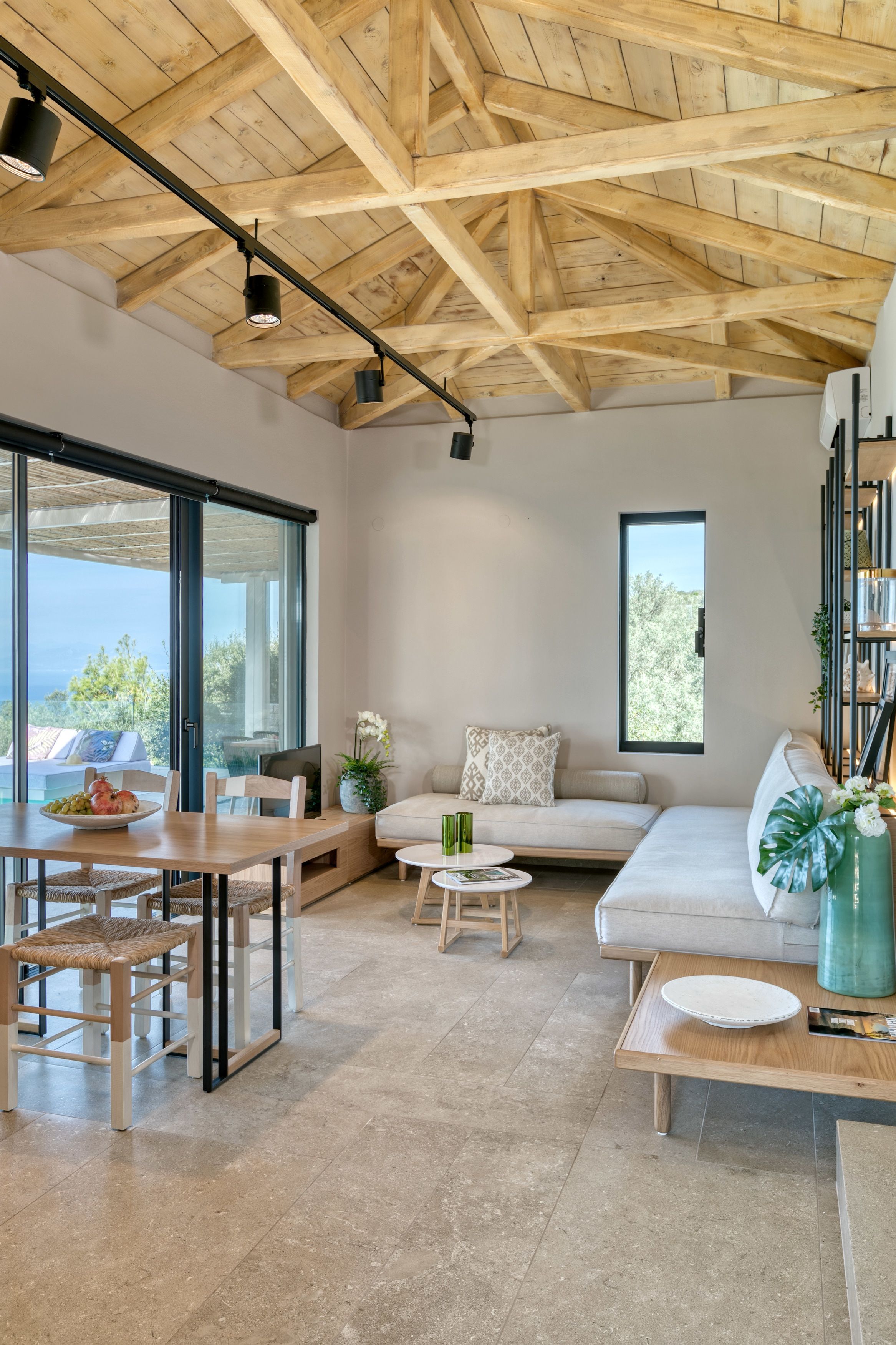 Amplia sala de estar con techo alto de madera, un gran ventanal corredera que da a una terraza exterior, un sofá, una mesa de centro y una mesa de comedor con sillas.