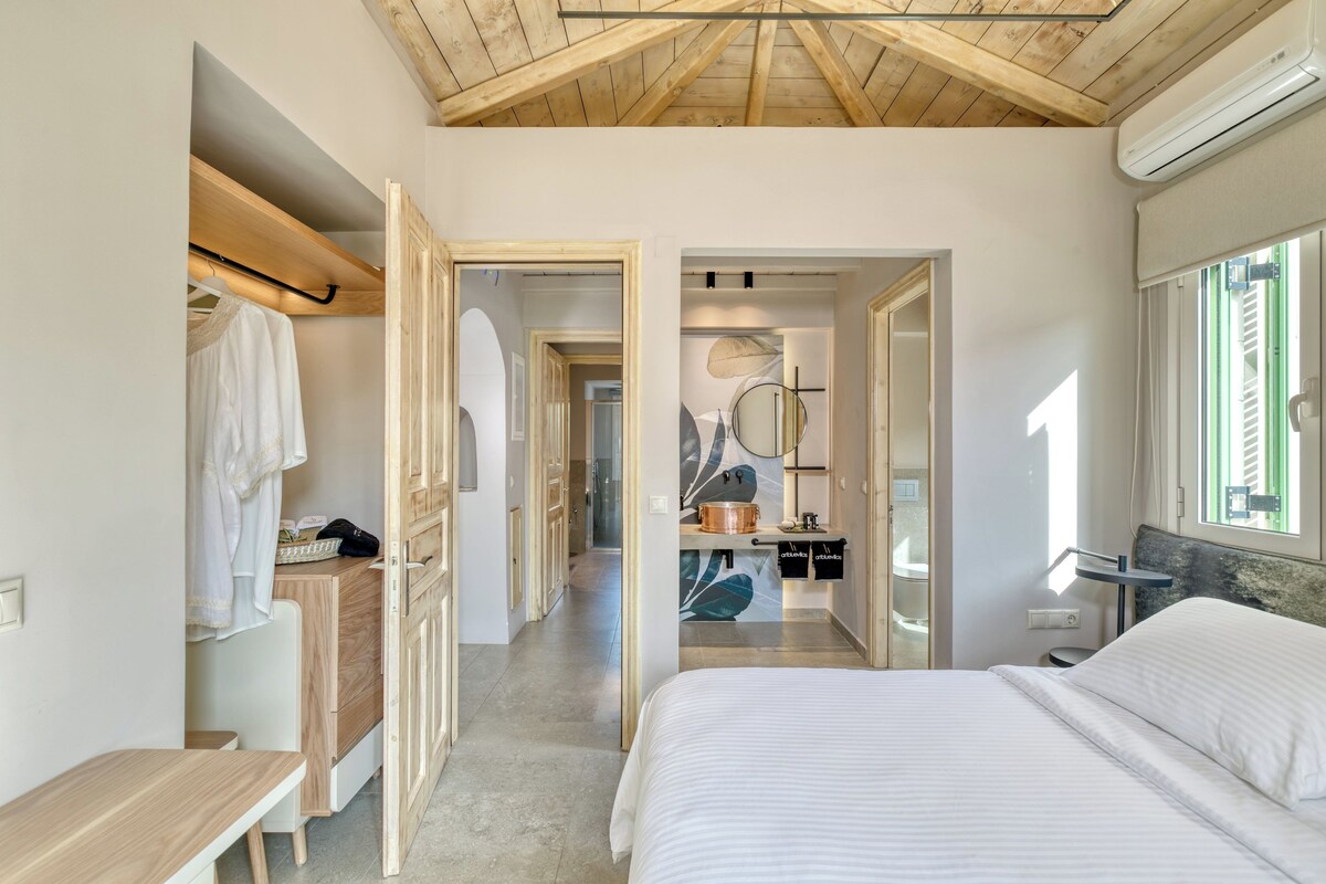 A bedroom with a large bed, wooden ceiling beams, a walk-in closet, and a bathroom with a shower and a mirror.