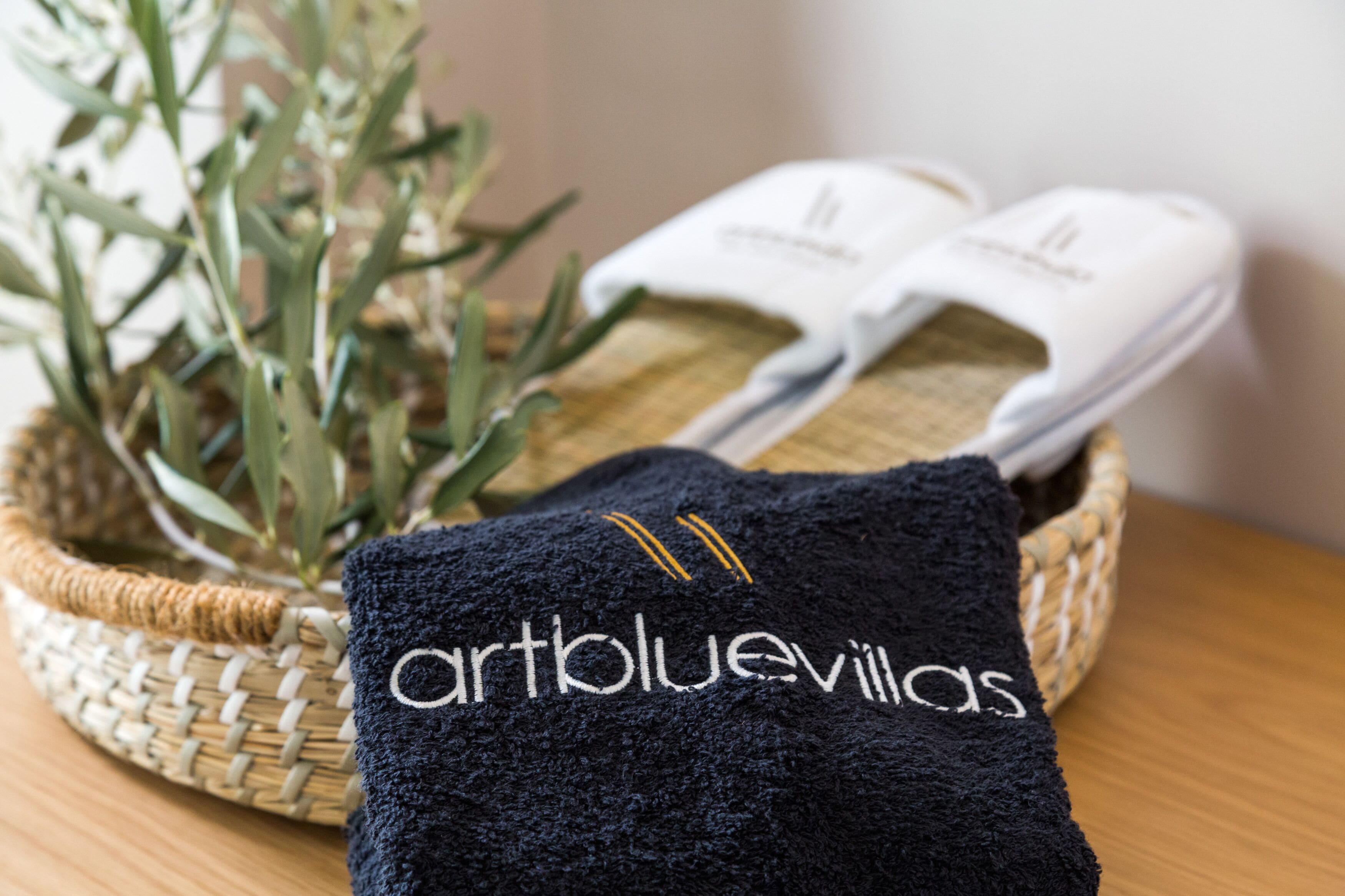 A woven basket containing a black towel with embroidered text, a pair of white slippers, and a potted plant.