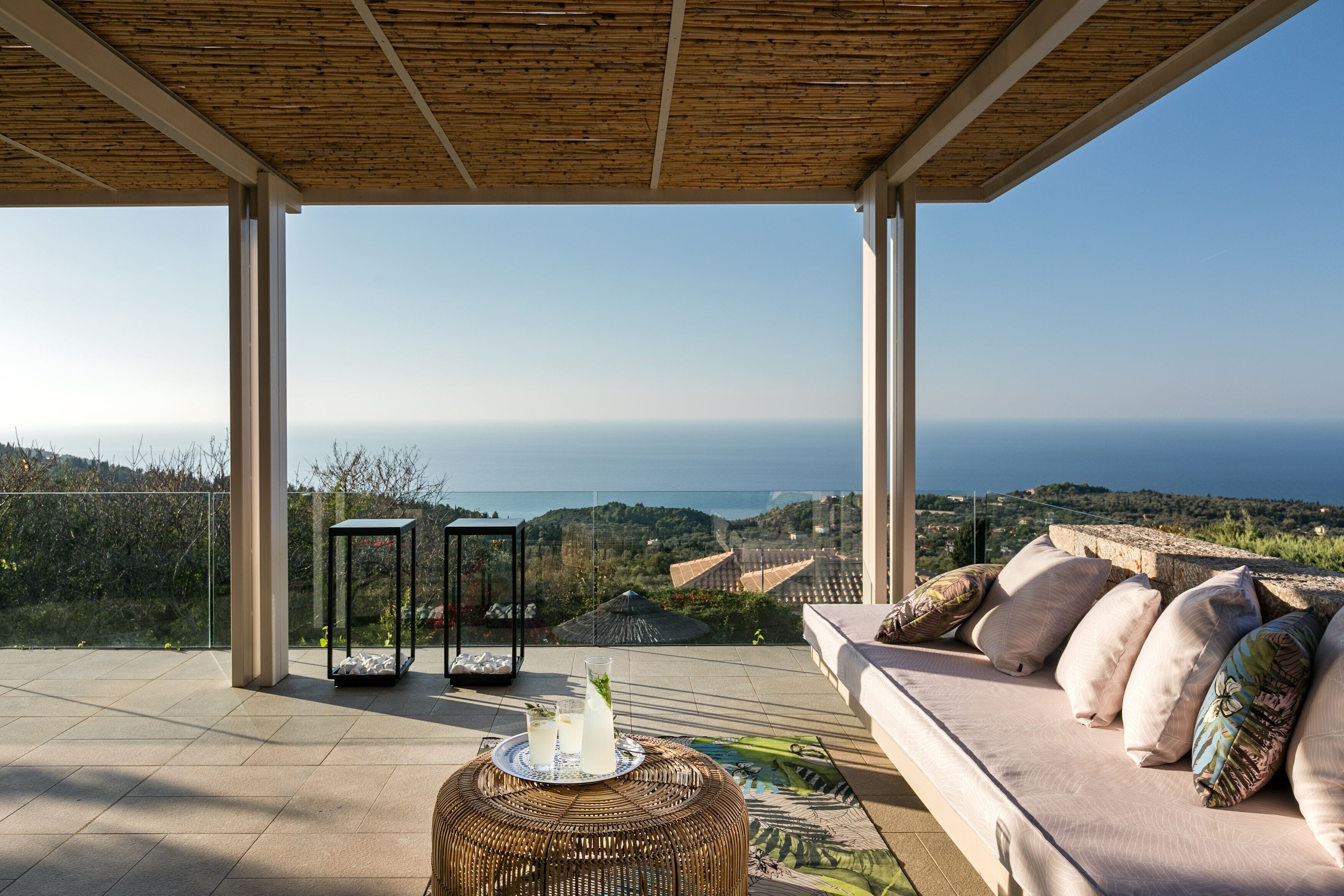 A modern outdoor seating area with a view of the sea, a wicker table with drinks, and a cushioned sofa.