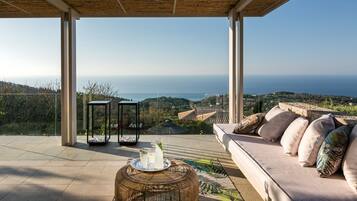 A modern outdoor seating area with a view of the sea, a wicker table with drinks, and a cushioned sofa.