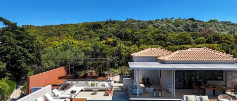 Una villa moderna con piscina, mobiliario exterior y vistas a una ladera boscosa.