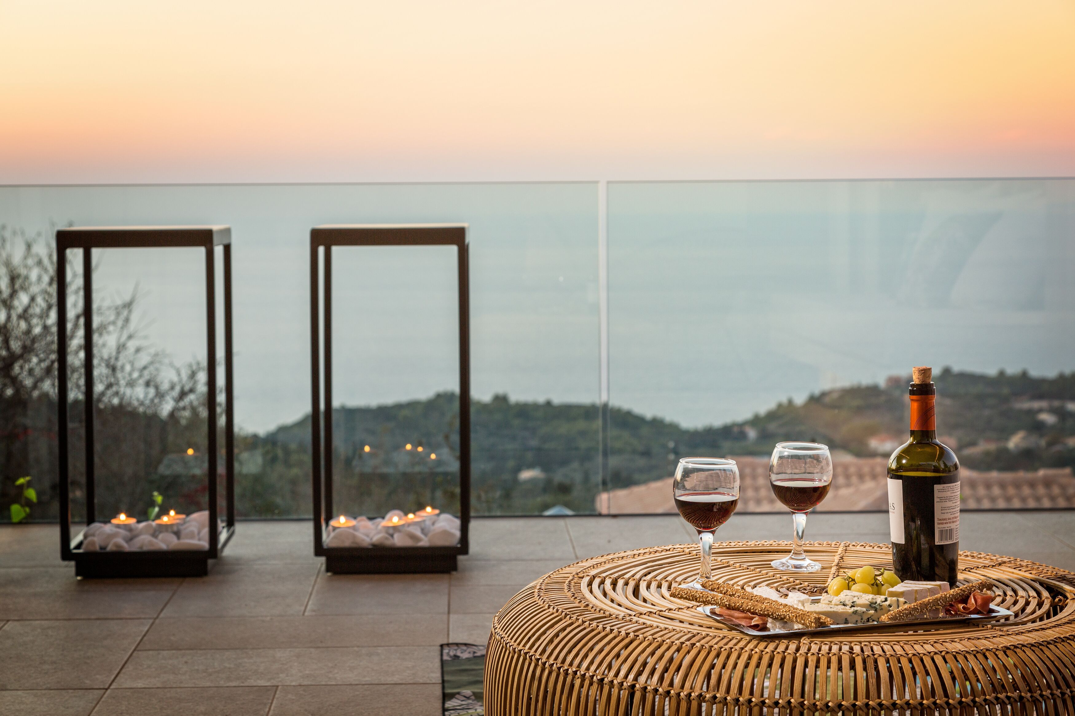 A terrace with a wicker table set for a meal, two glasses of red wine, a bottle of wine, and candles in two glass lanterns.