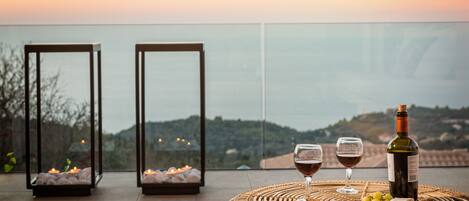 A terrace with a wicker table set for a meal, two glasses of red wine, a bottle of wine, and candles in two glass lanterns.