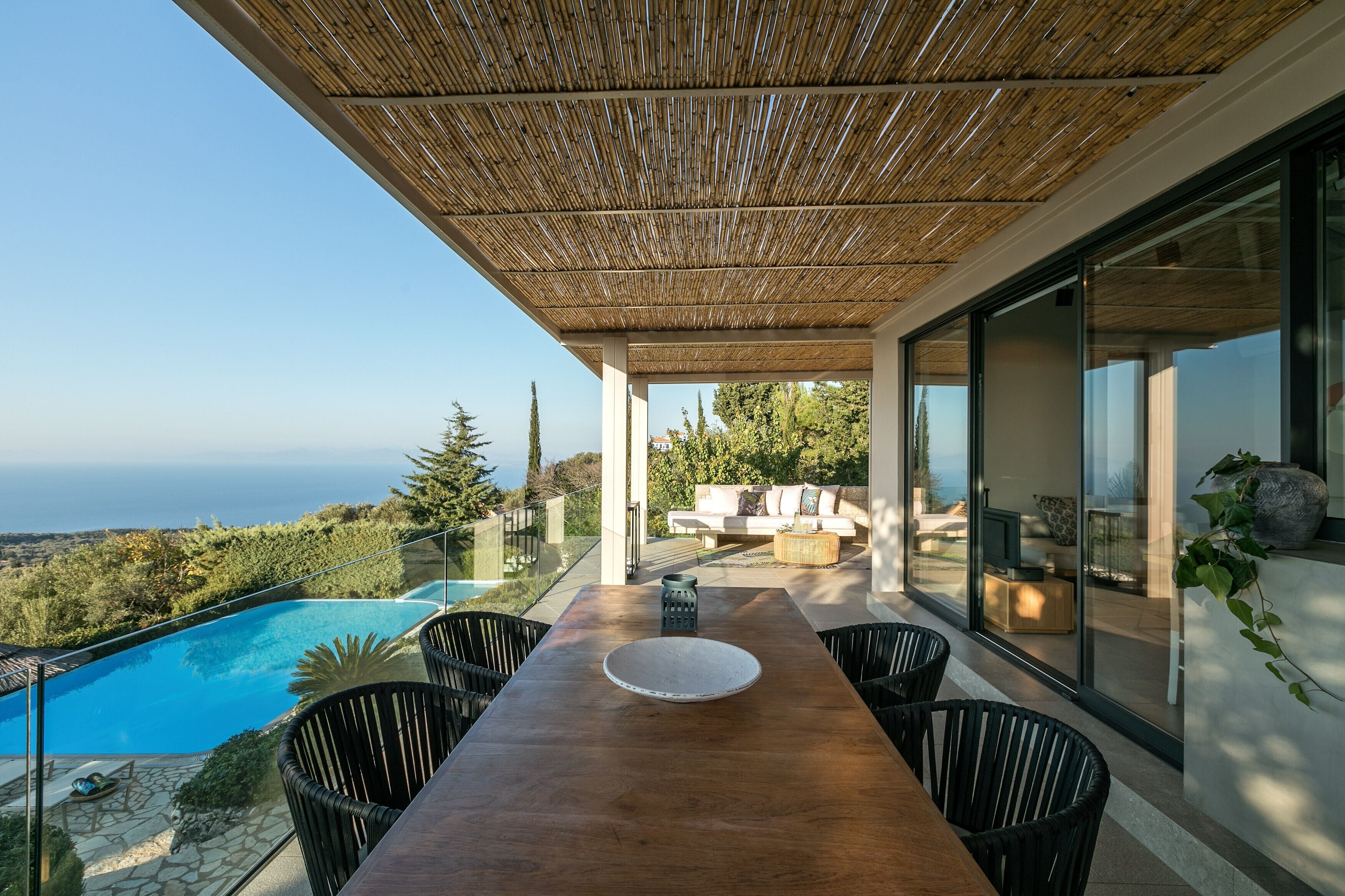 Un espace repas extérieur moderne, avec une table et des chaises en bois, offrant une vue sur une piscine et un paysage marin pittoresque.