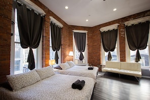 Quarto de hotel com duas camas, um sofá e amplas janelas com cortinas.