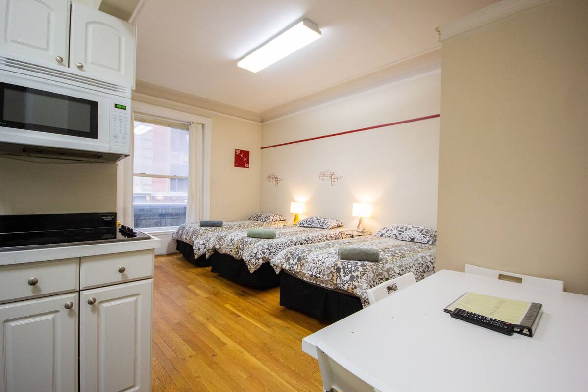 A hotel room with a kitchenette, two beds with floral bedding, bedside lamps, and a white table with a remote control.
