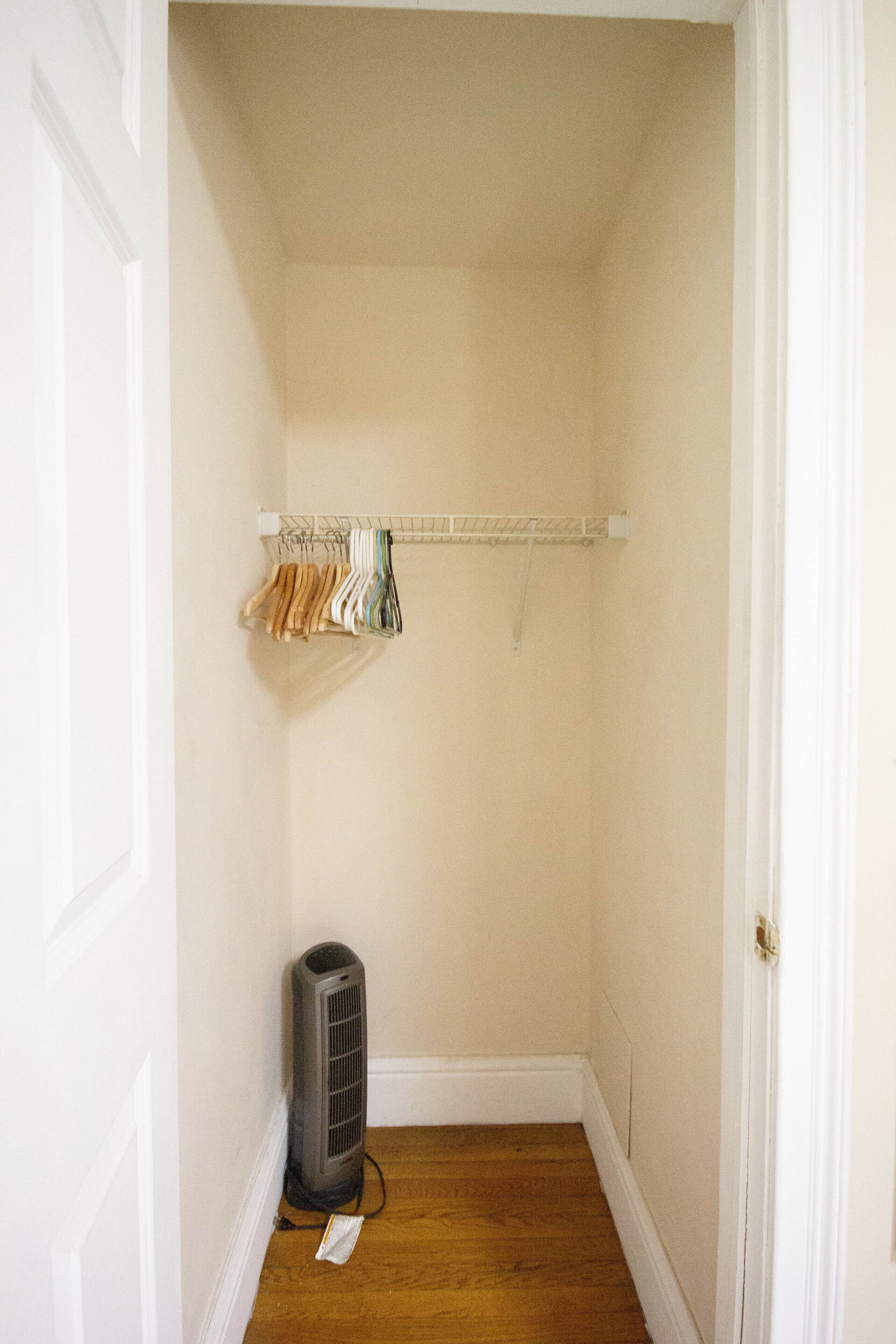 A small, narrow room with a wooden floor, a white door, a closet with hanging clothes, and a standing heater.