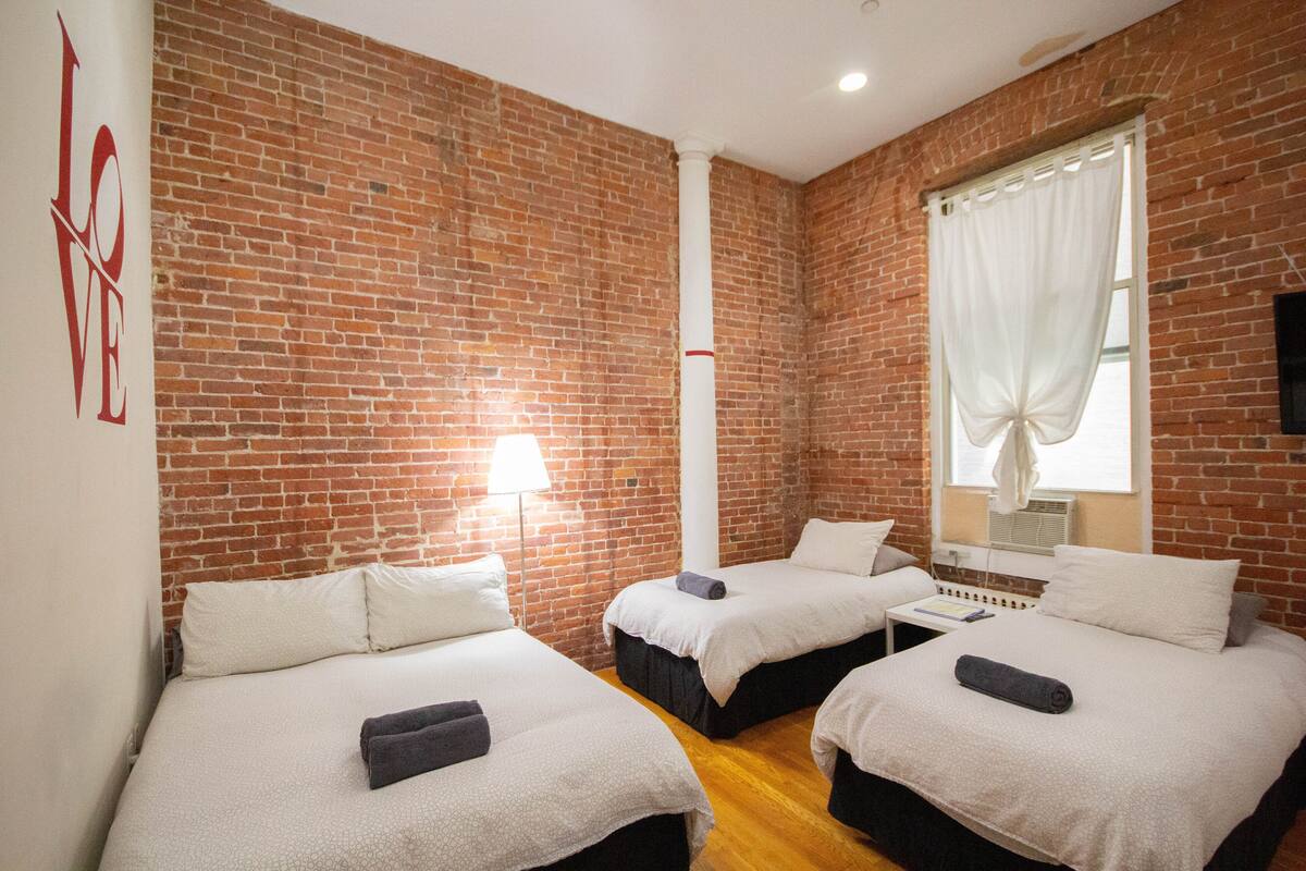 A hotel room with two beds, a brick wall, a window with white curtains, and a wall-mounted TV.