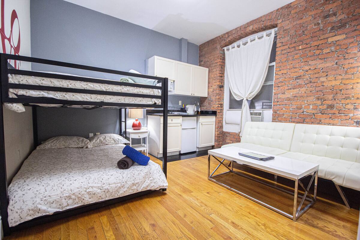 A room with a bunk bed, a white sofa, a small table, and a kitchen area with cabinets and a refrigerator.