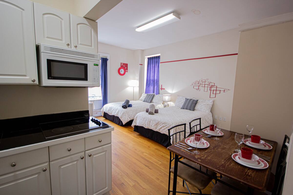 A compact hotel room with a kitchenette, two beds, a dining table set for four, and a wall clock.