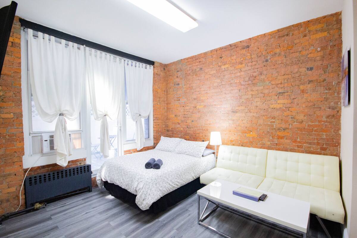 A room with a brick wall, a bed with a white duvet, a white sofa, and a small table with a remote control.