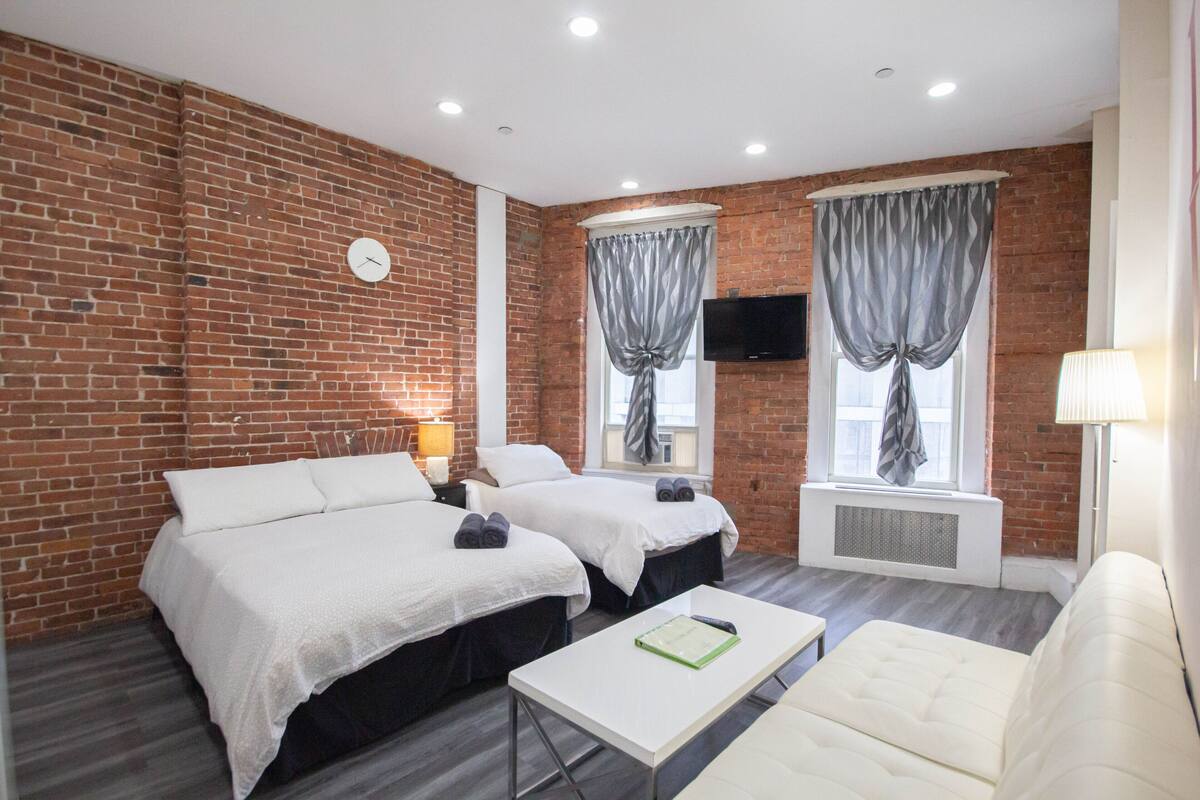 A hotel room with two beds, a white sofa, a small table, a TV, and brick walls.