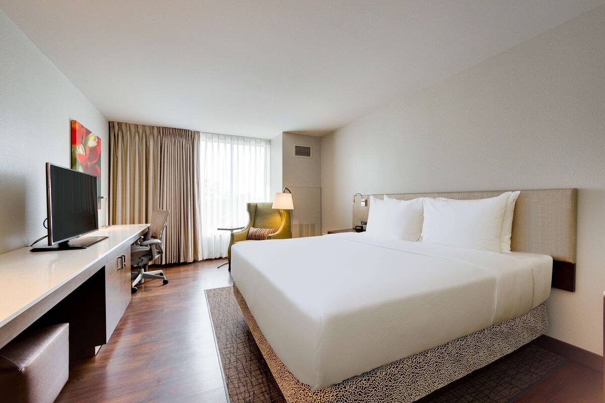 A hotel room with a large bed, a desk with a computer, a chair, a lamp, and a window with curtains.