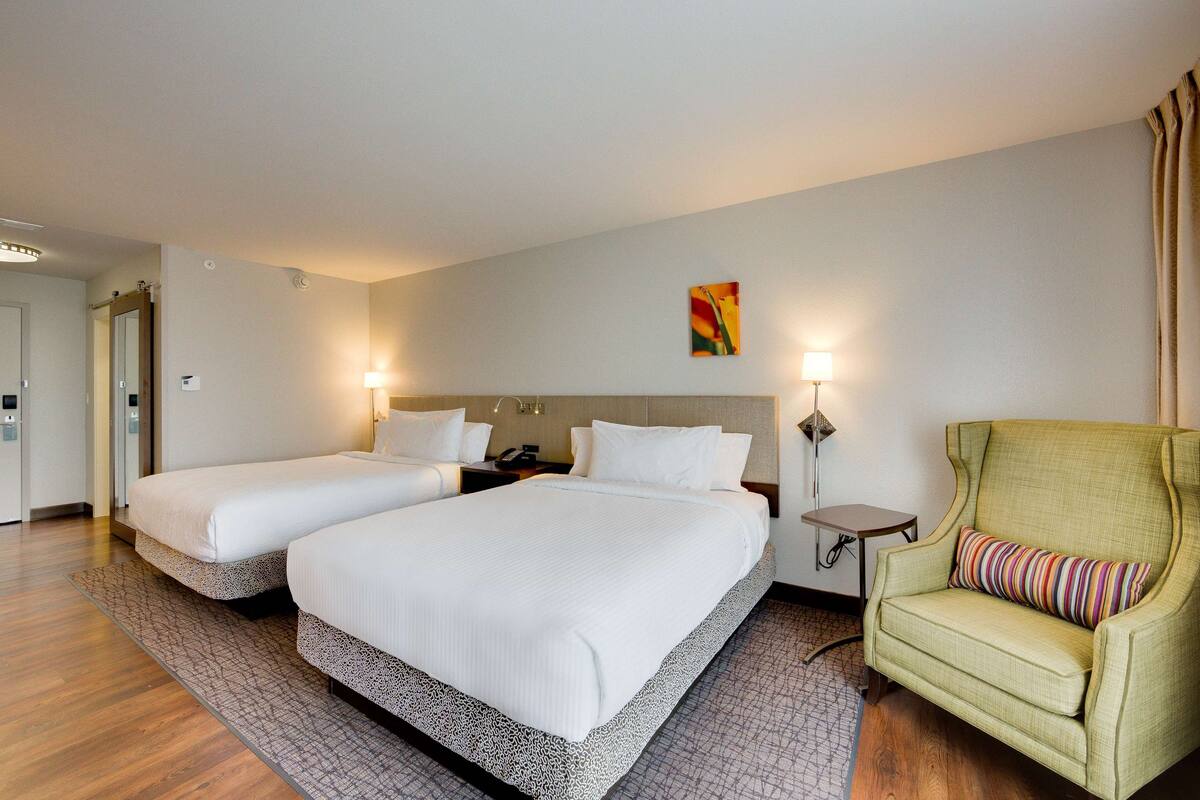 A hotel room with two beds, a green armchair, and a small table with a lamp.