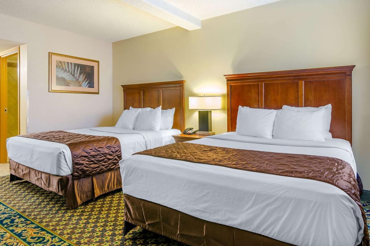 A hotel room with two beds, a patterned carpet, a painting on the wall, and a nightstand with a lamp.