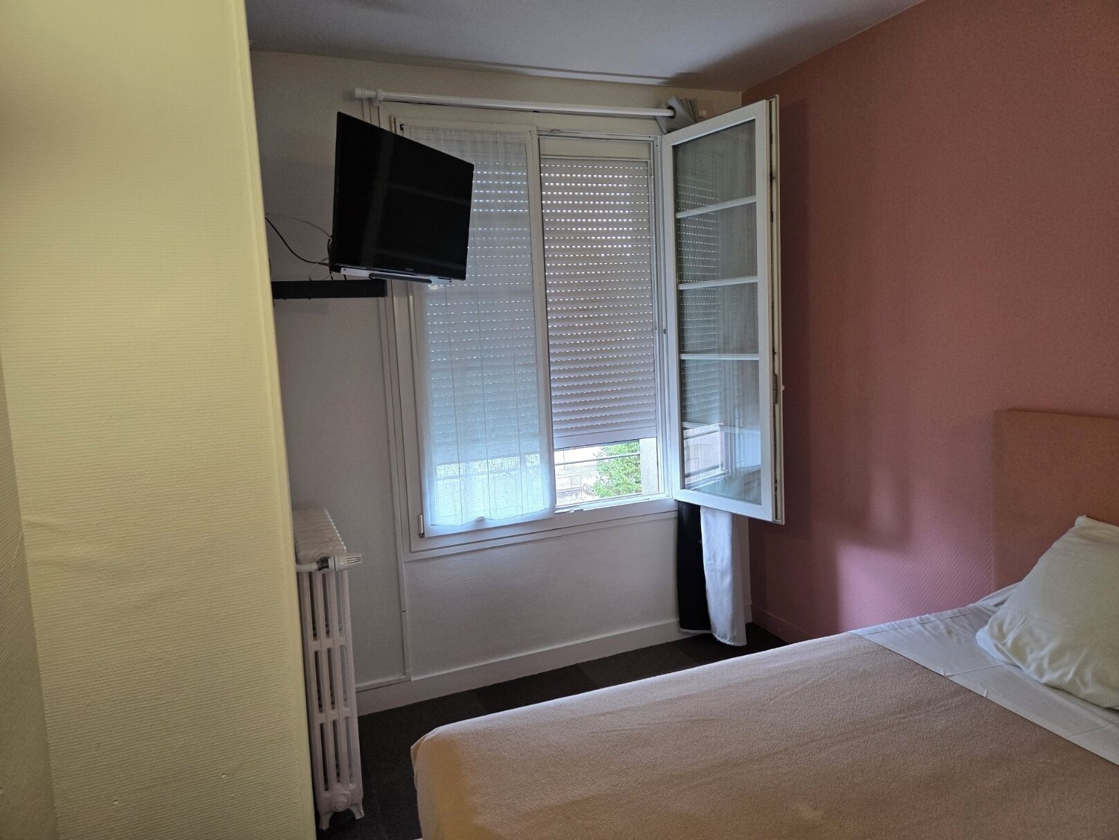A hotel room with a bed, a television mounted on the wall, and a window with blinds.