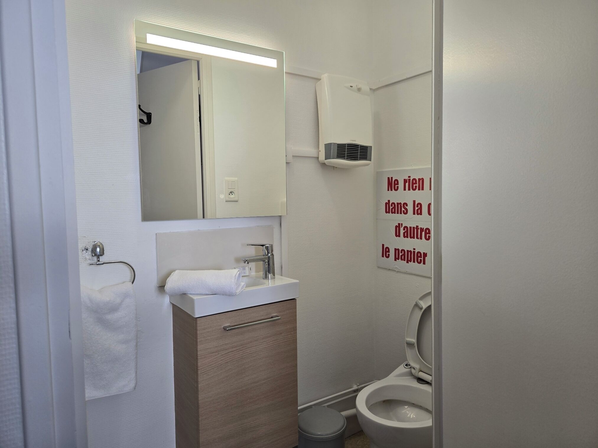 Un baño con lavamanos, espejo, inodoro y un cartel en la pared.