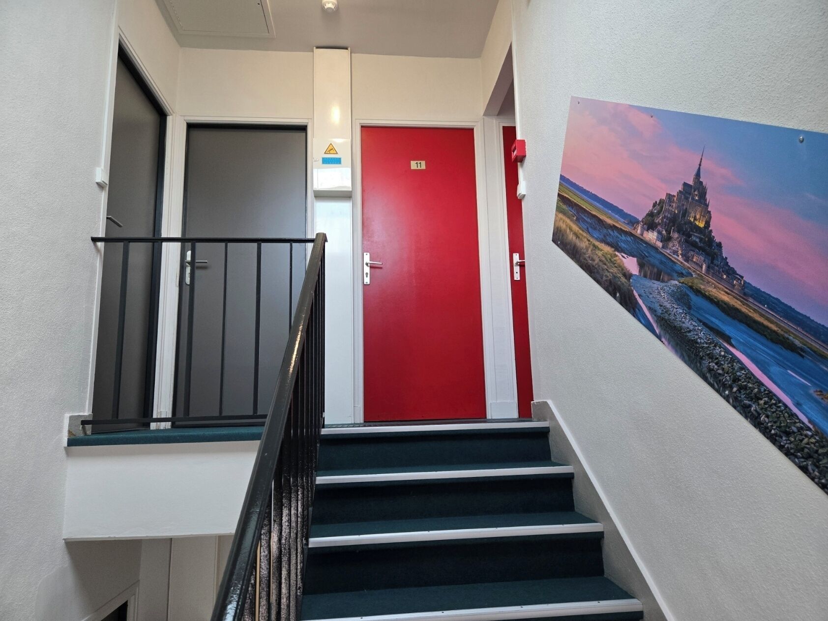 Una escalera con una puerta roja al final, una barandilla negra y un cuadro de un castillo en la pared.