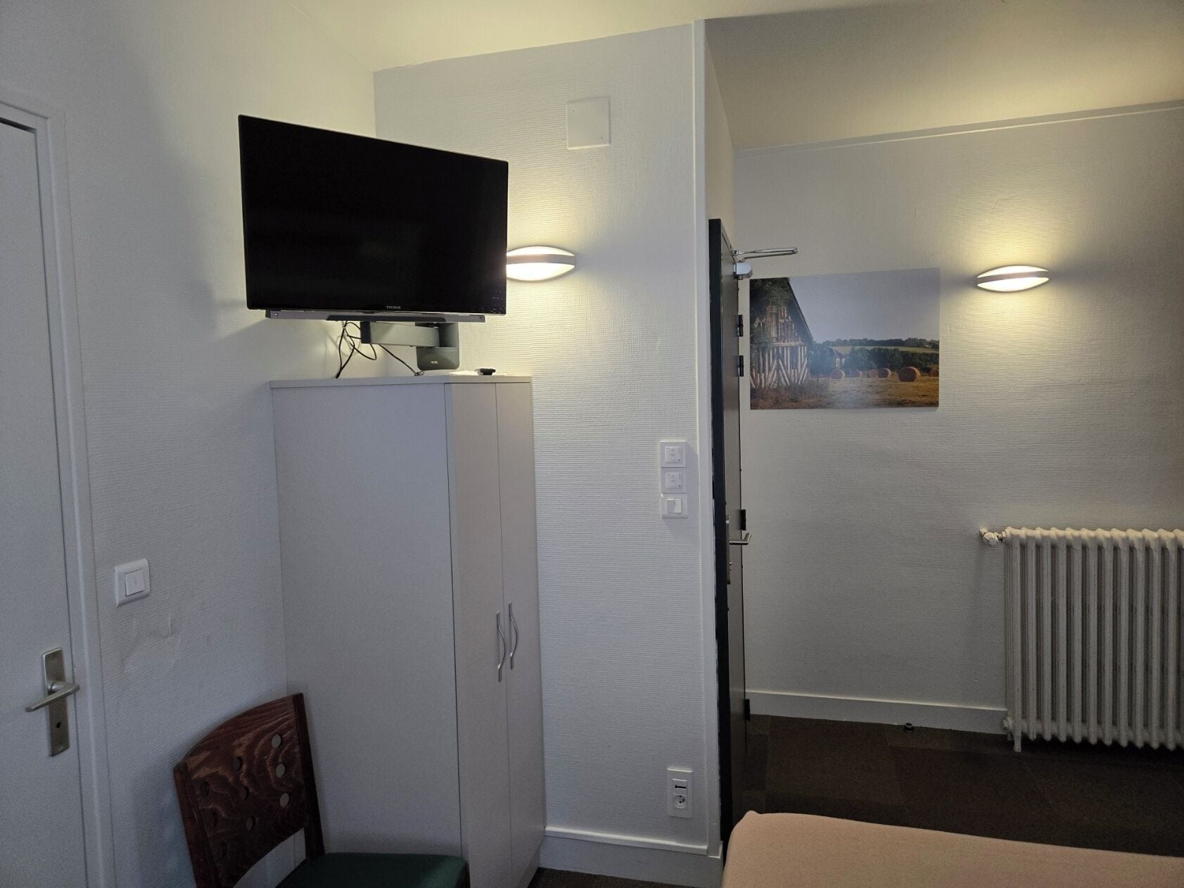 A room with a television mounted on a cabinet, a chair, a radiator, and a wall-mounted light fixture.