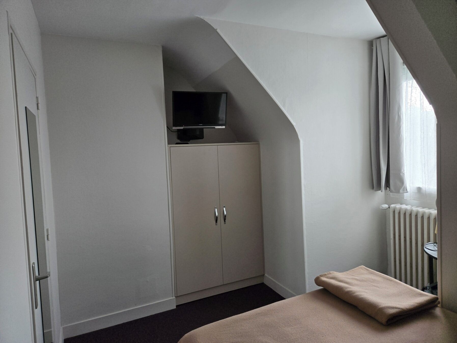 Un dormitorio con una televisión sobre un mueble, un radiador y una ventana con cortinas.