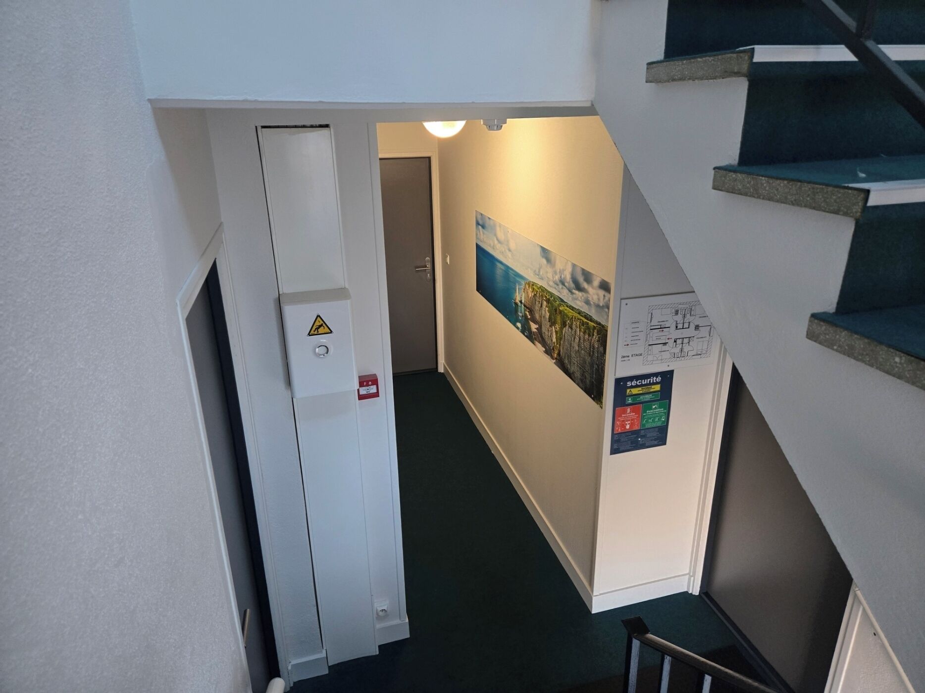 Un pasillo con una escalera, una puerta con detector de incendios y un cartel de seguridad.