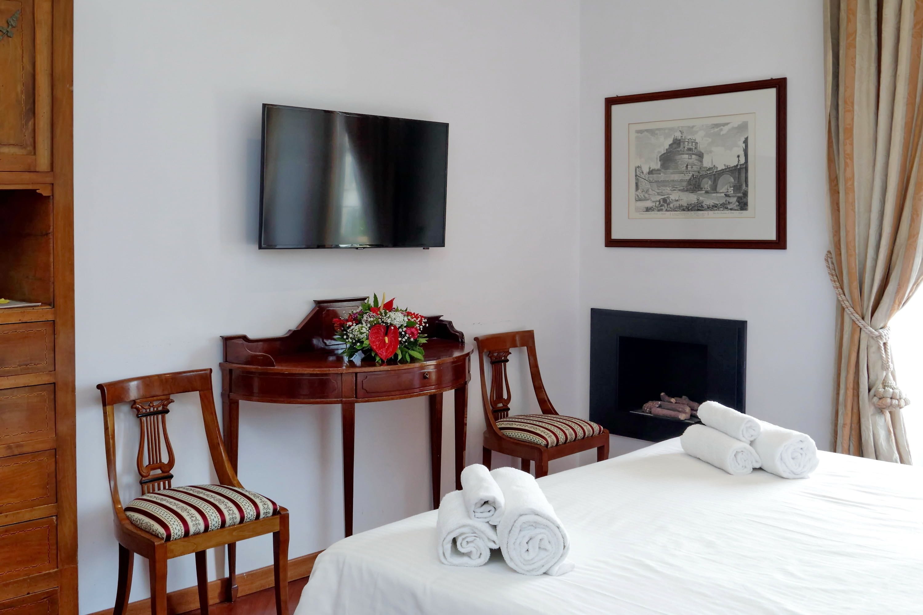 A hotel room with a bed, two wooden chairs, a small table with a vase of flowers, a flat-screen TV mounted on the wall, and a framed picture above the fireplace.