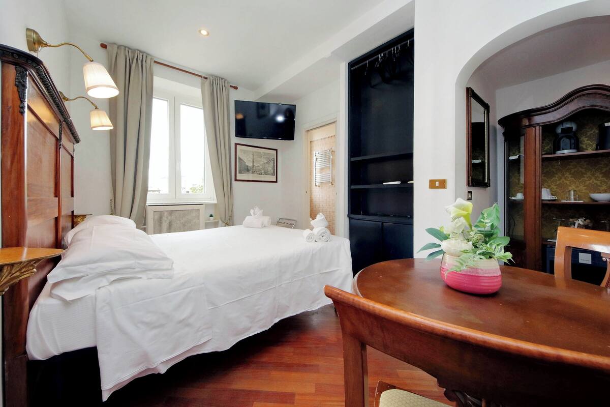 A hotel room with a bed, a desk, a chair, a TV, a bookshelf, and a window with curtains.