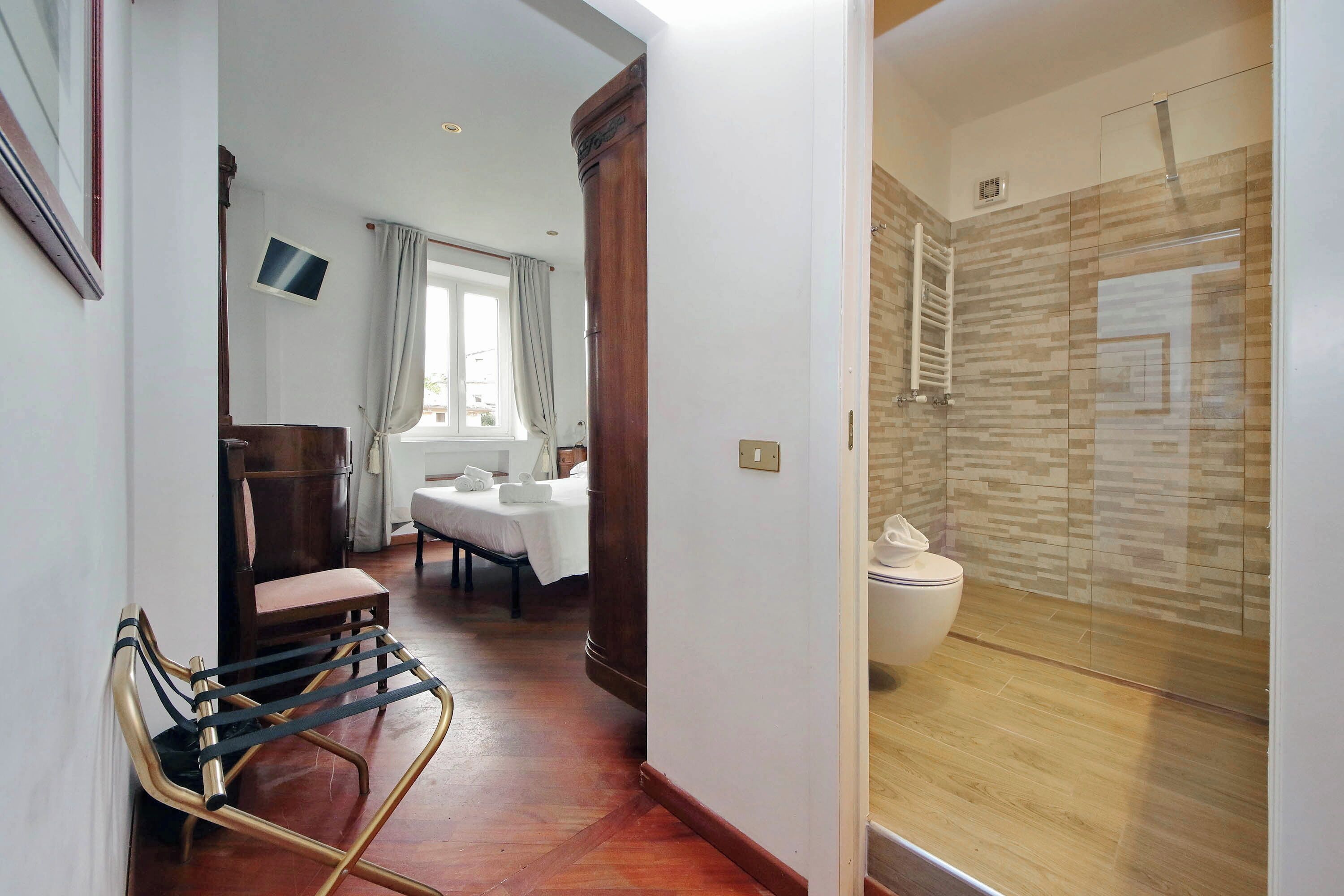 A hotel room with a wooden floor, a bed with a white duvet, a wooden chair, a small table, a window with curtains, and a shower area with a glass door.