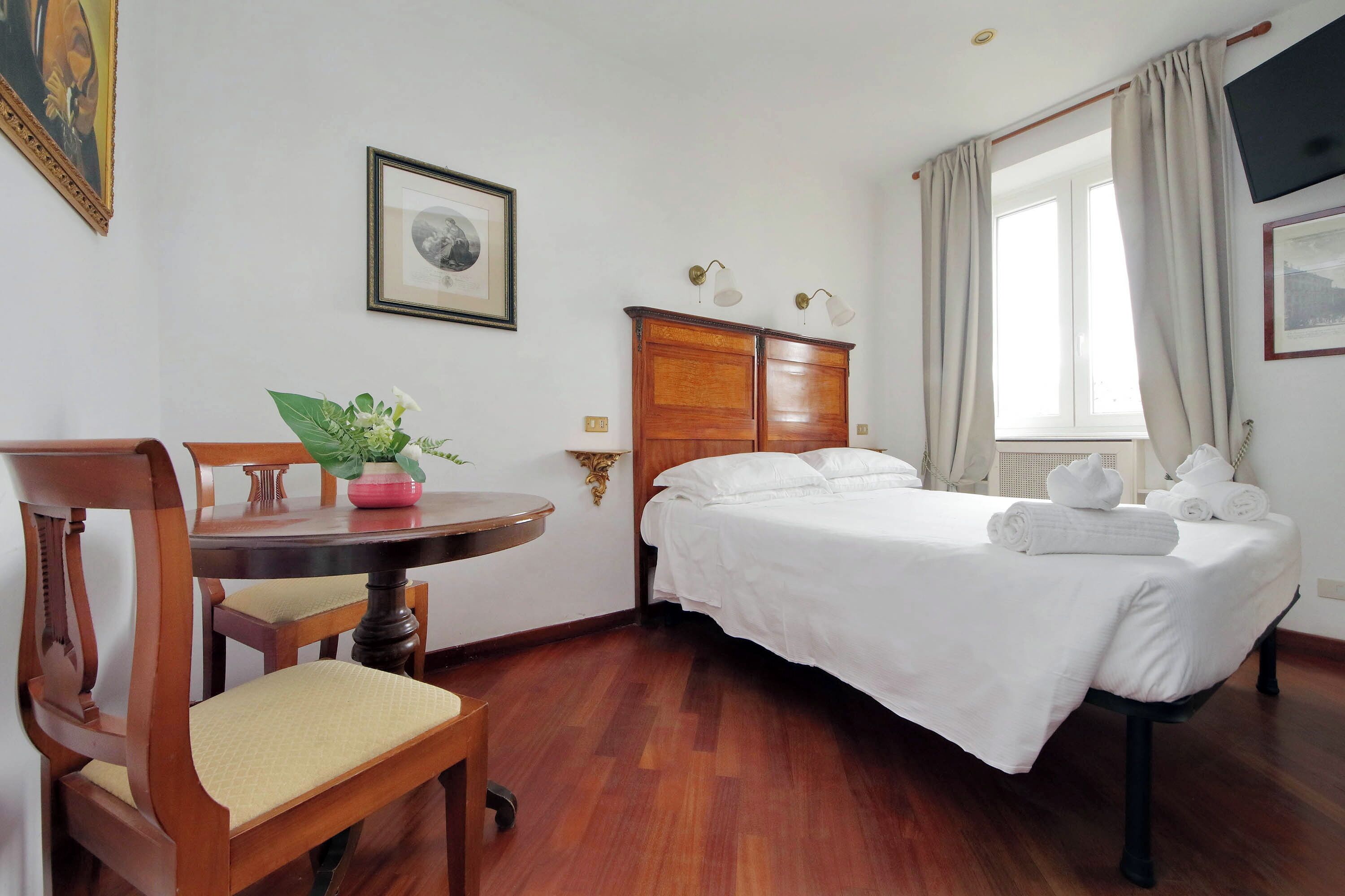 Een hotelkamer met een bed, een houten stoel, een klein tafeltje met een plant, een lijst met foto en een raam met gordijnen.