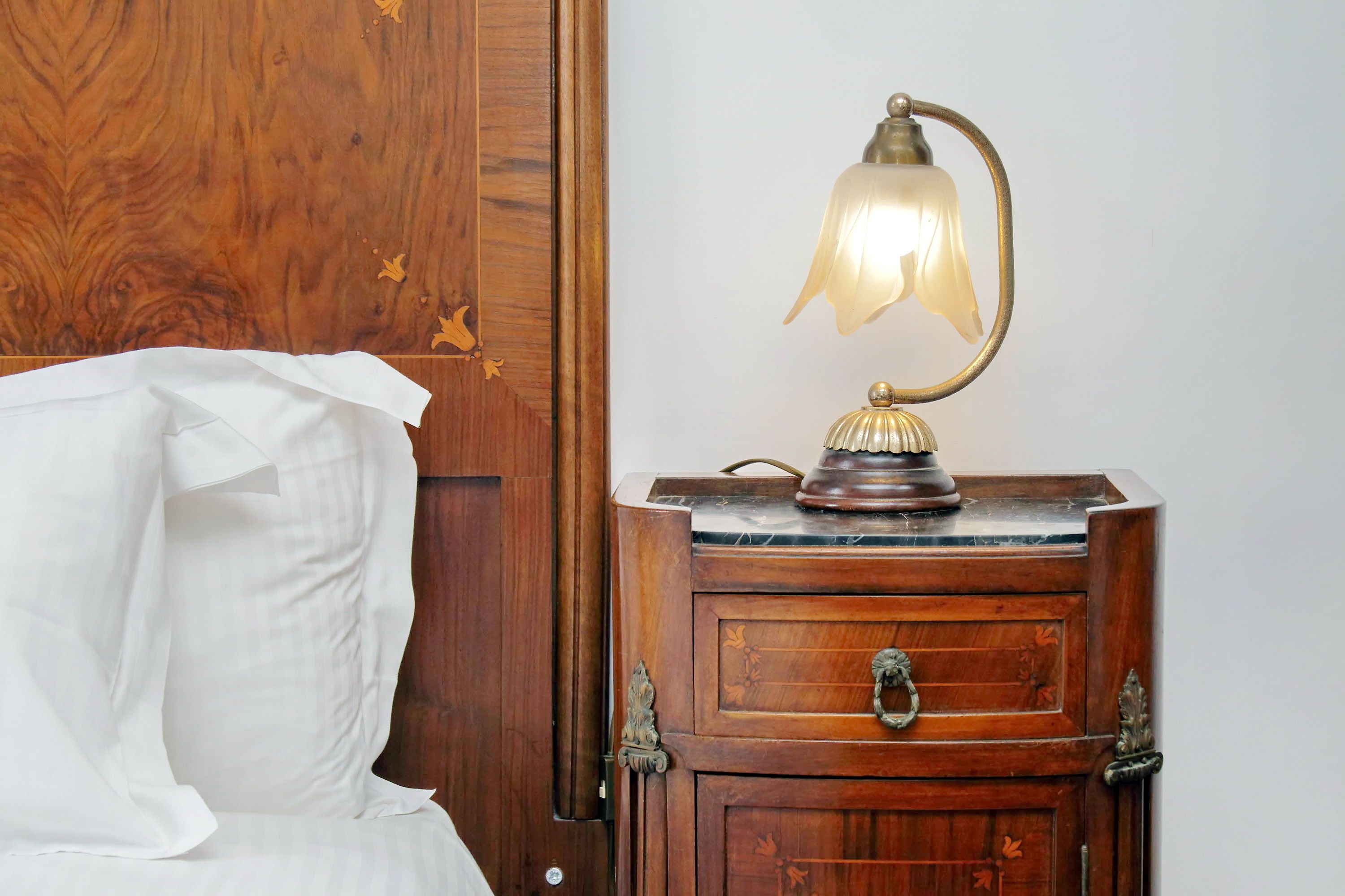 A wooden nightstand with a lamp, a drawer, and a bed with a white sheet.
