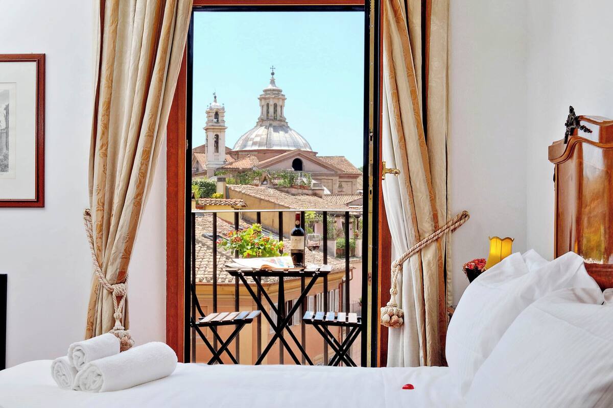 A hotel room with a bed, a small table with chairs, a view of a church, and a balcony with a rope.