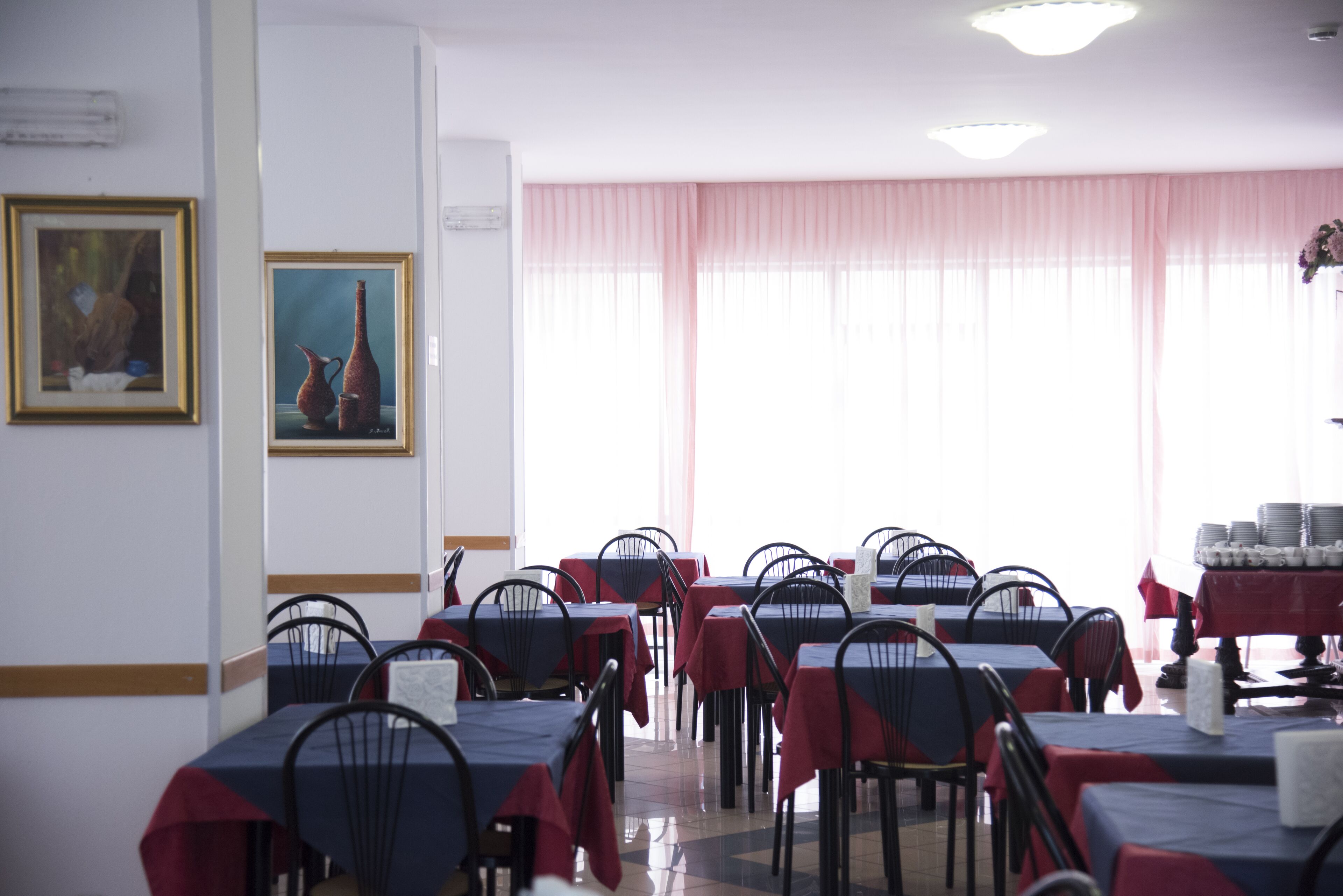 A dining area with tables and chairs, framed artwork on the walls, and a large window with sheer curtains.