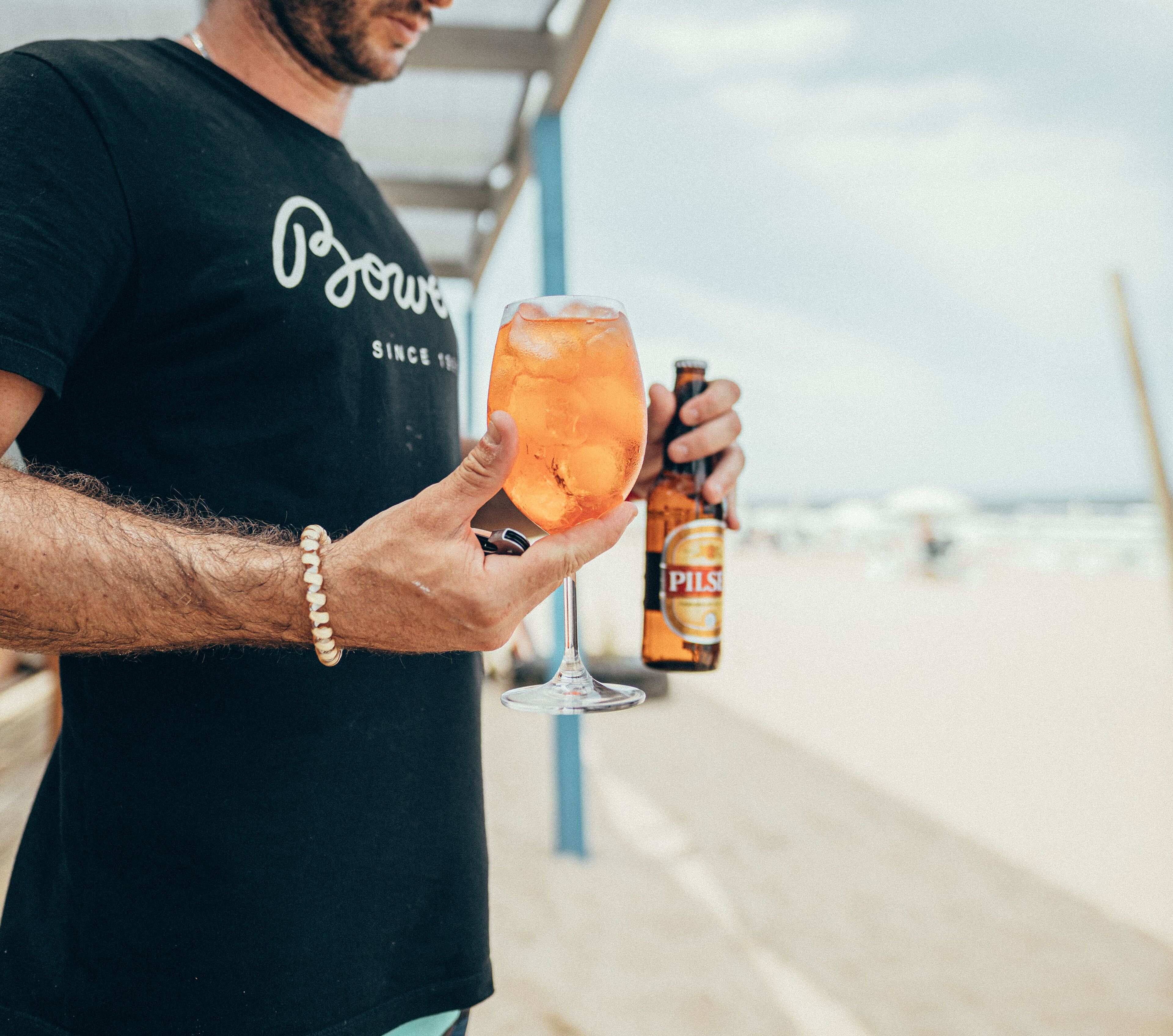 A person holding a glass of beer and a bottle of Pilsner beer on a beach.