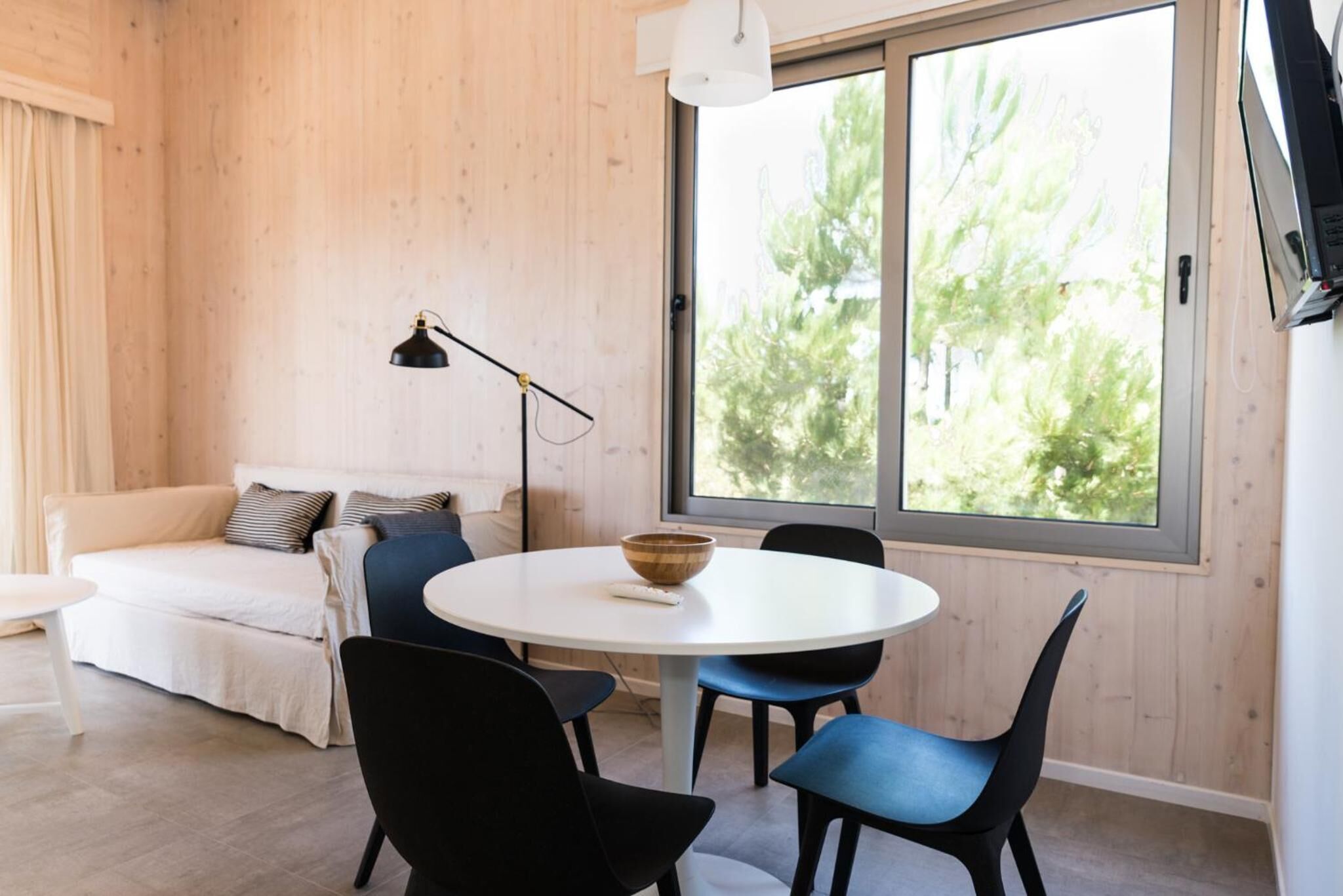 A cozy room with a round white table, blue chairs, a white sofa, and a large window with a view of trees.