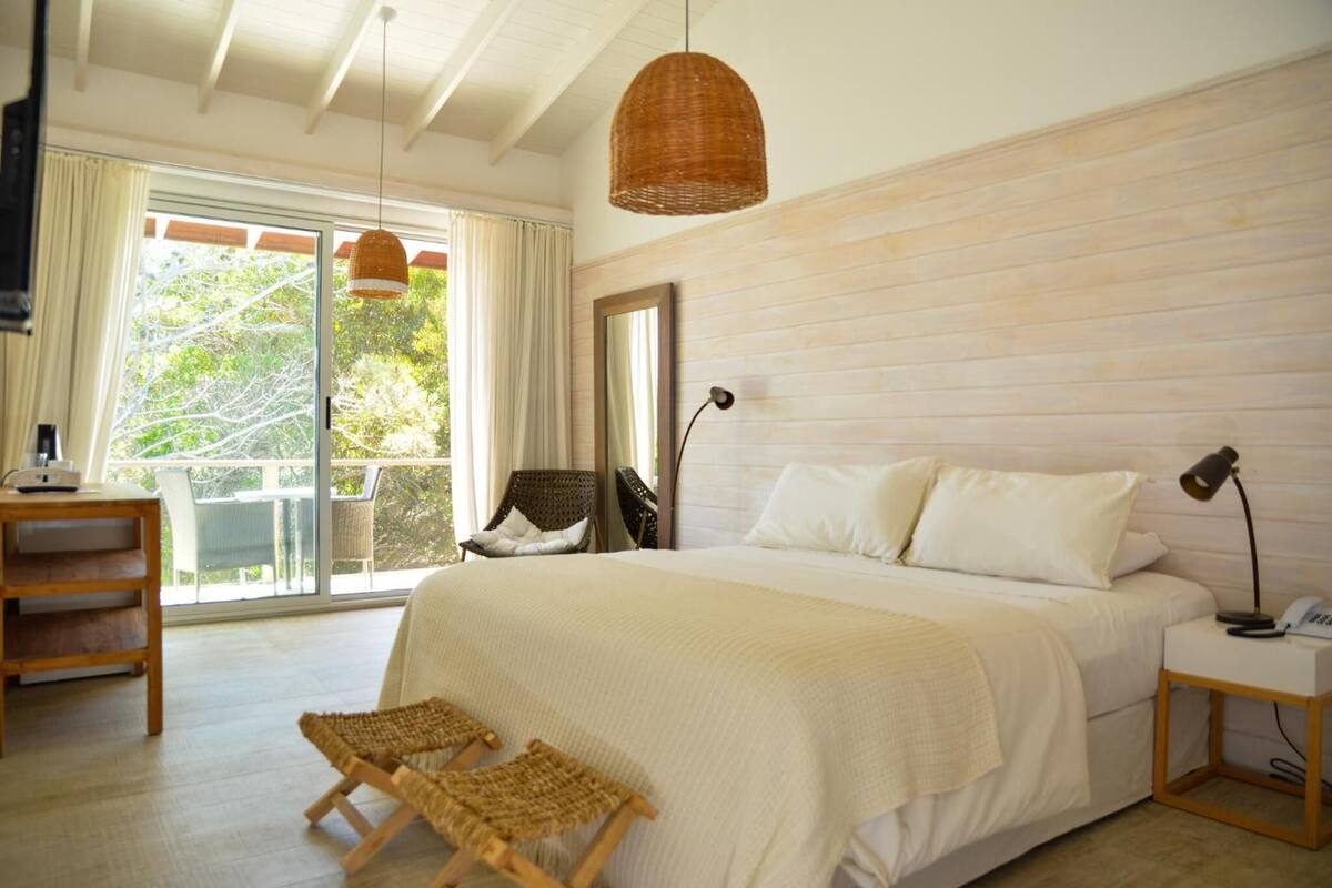 A bedroom with a large bed, a desk, wicker chairs, a hanging light, and a view of greenery through the window.