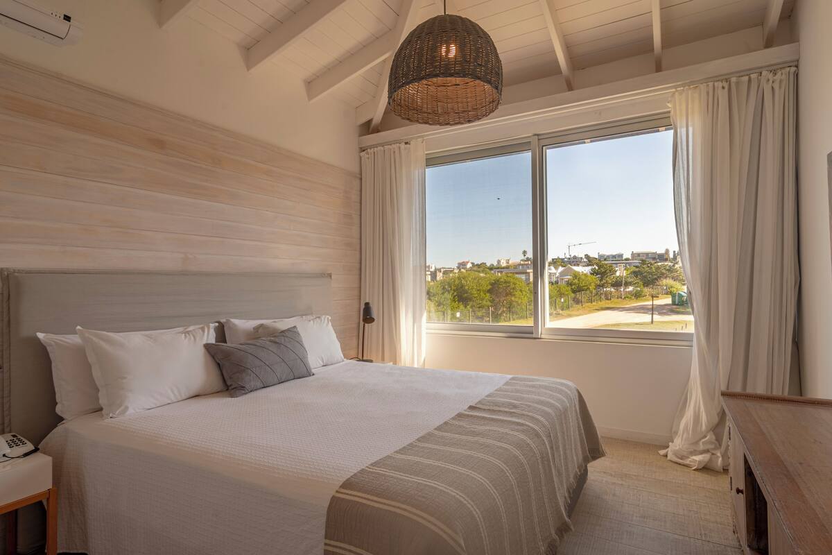 A bedroom with a large bed, a bedside table with a phone, a window with curtains, and a view of the city.